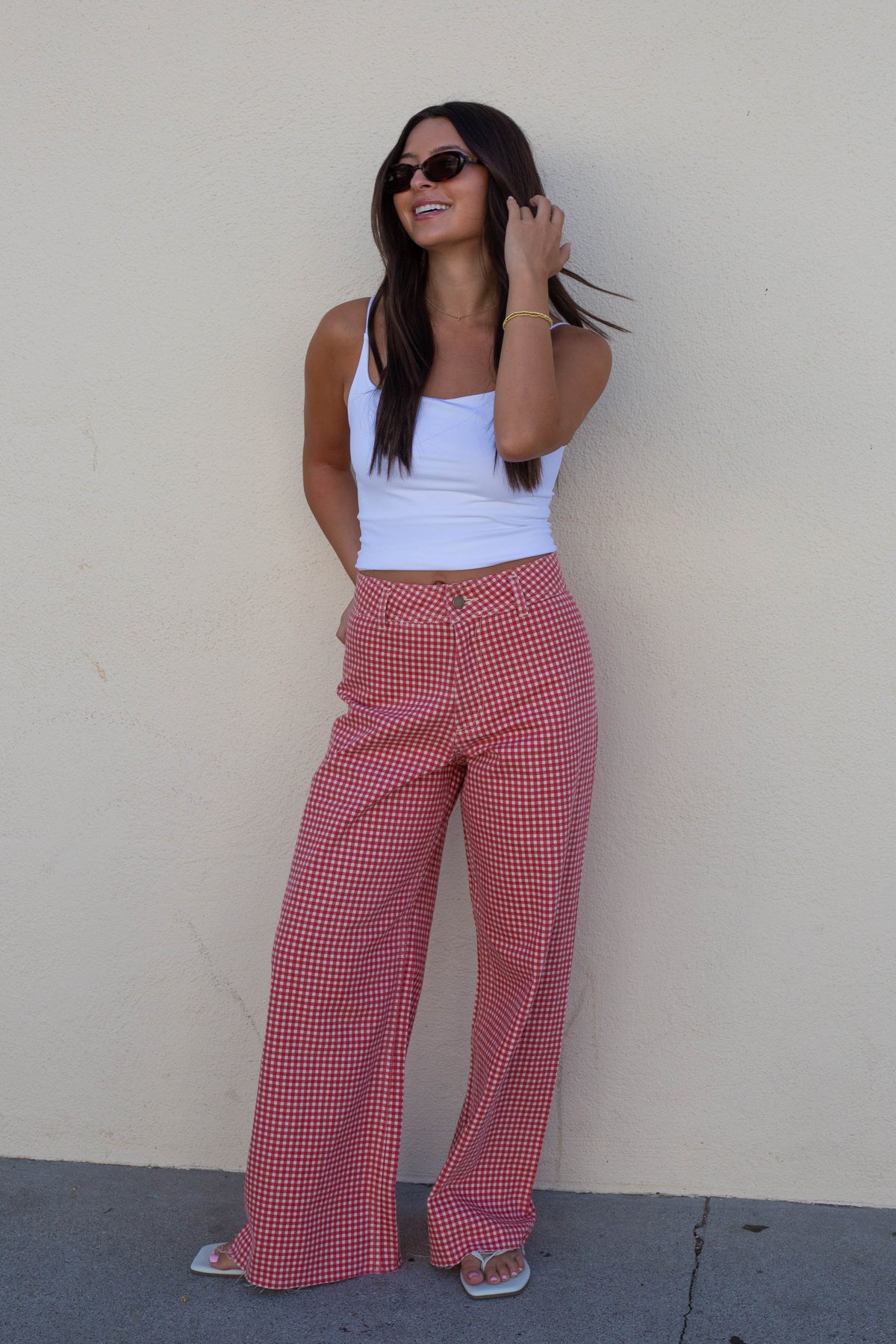A woman wearing red and white gingham wide-leg pants, a white tank top, and sunglasses stands against a plain beige wall.