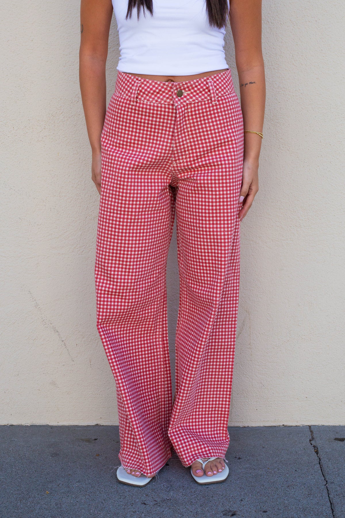 A person wearing red and white gingham wide-leg pants, a white tank top, and white slide sandals, standing against a beige stucco wall.