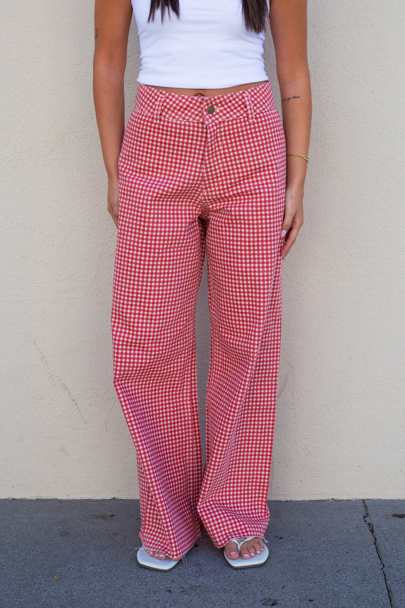 A person wearing red and white gingham wide-leg pants, a white tank top, and white slide sandals, standing against a beige stucco wall.
