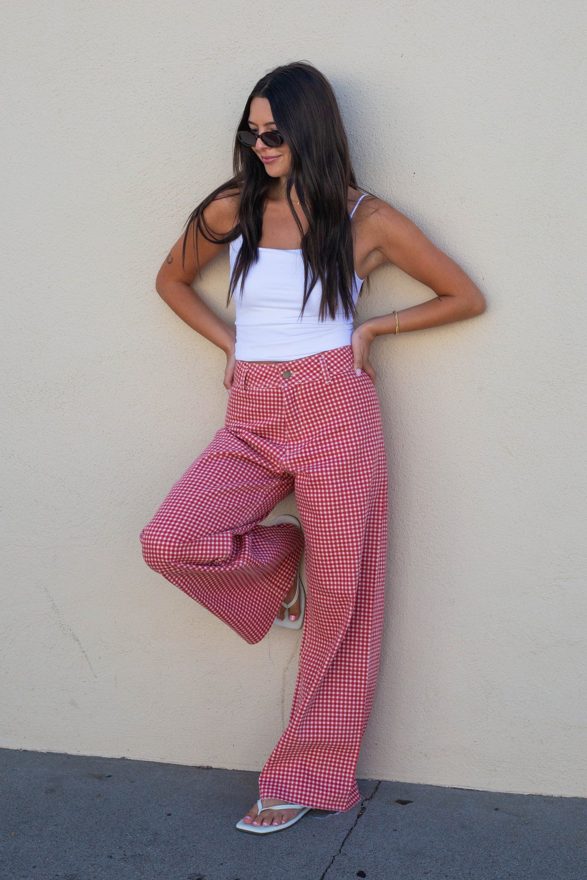 A woman wearing red and white gingham wide-leg pants, a white tank top, and sunglasses, posing against a beige stucco wall.