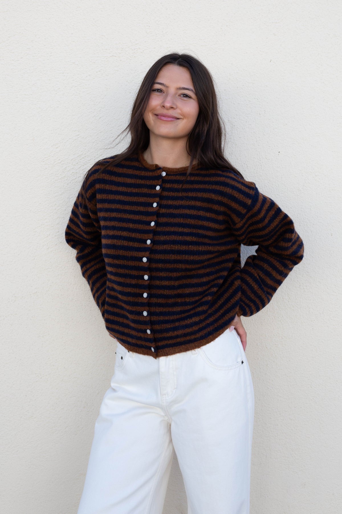 A woman wearing a dark brown and black striped knit cardigan with white buttons, paired with white jeans, standing against a textured off-white wall.