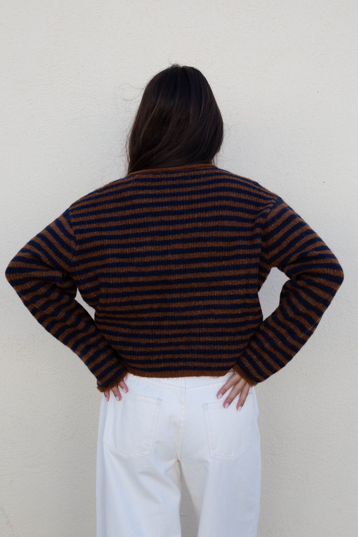 A person shown from the back wearing a dark brown and black horizontally striped knit sweater with white pants, standing against a plain off-white wall.