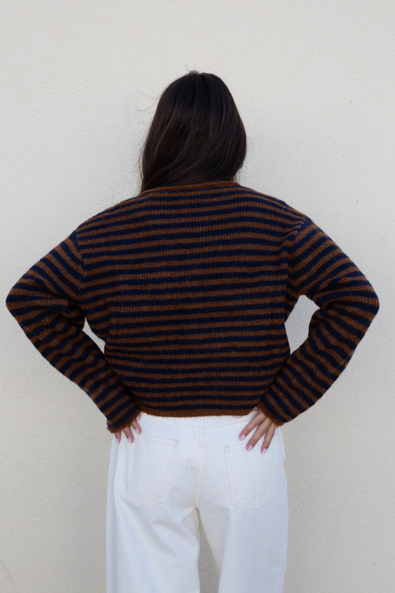 A person shown from the back wearing a dark brown and black horizontally striped knit sweater with white pants, standing against a plain off-white wall.