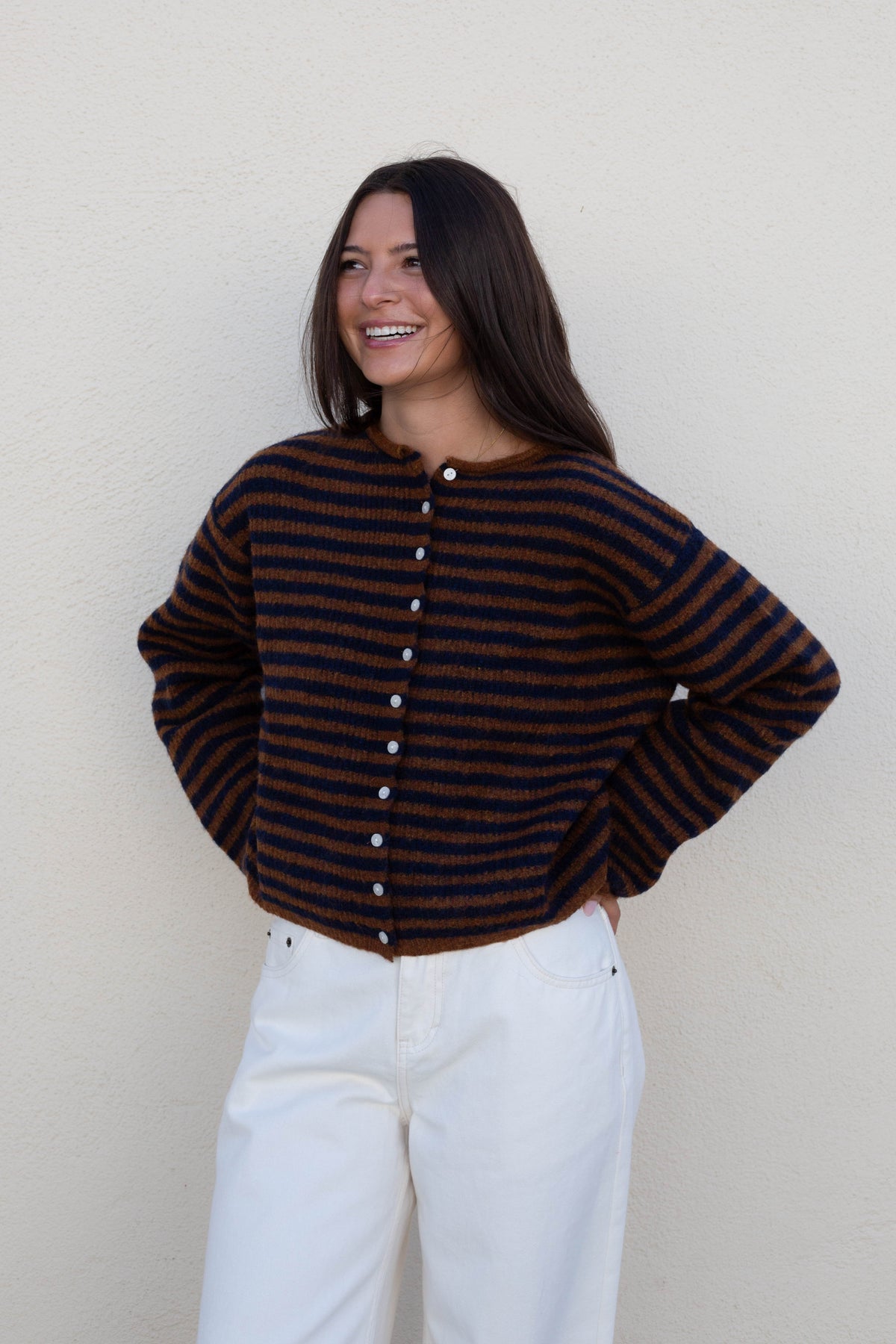 A woman wearing a dark brown and black striped knit cardigan sweater with white buttons down the front, paired with white pants, standing against a textured off-white wall.