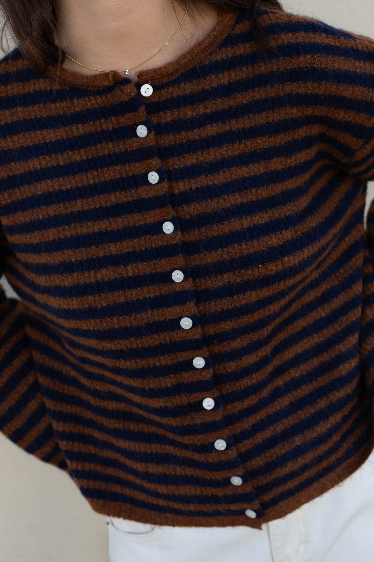 A close-up of a person wearing a brown and navy blue striped knit cardigan with white buttons, paired with white pants.