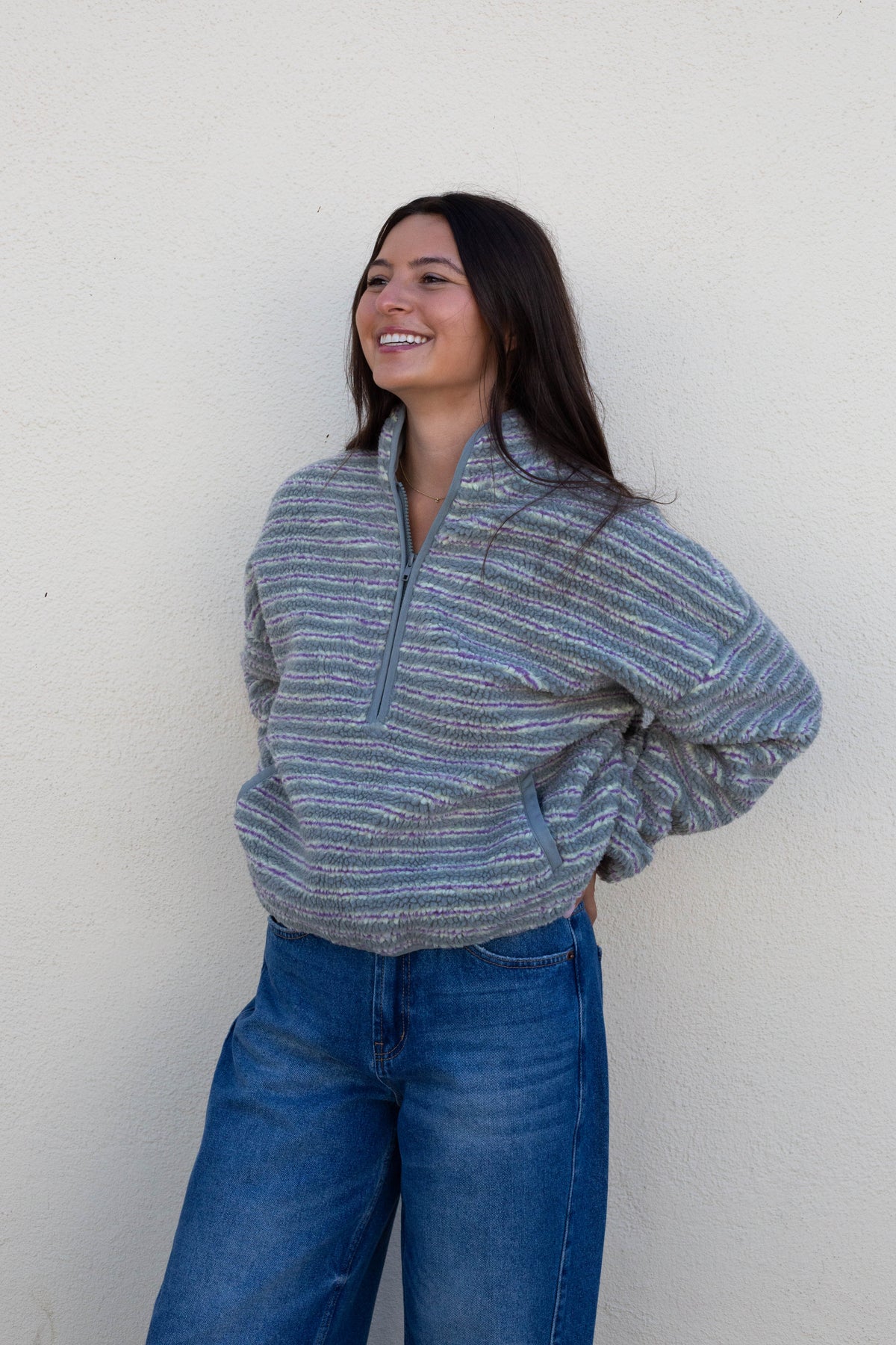 A woman with long dark hair smiling and looking to the side, wearing a gray and blue striped quarter-zip pullover sweater and blue jeans, standing against a plain off-white wall.