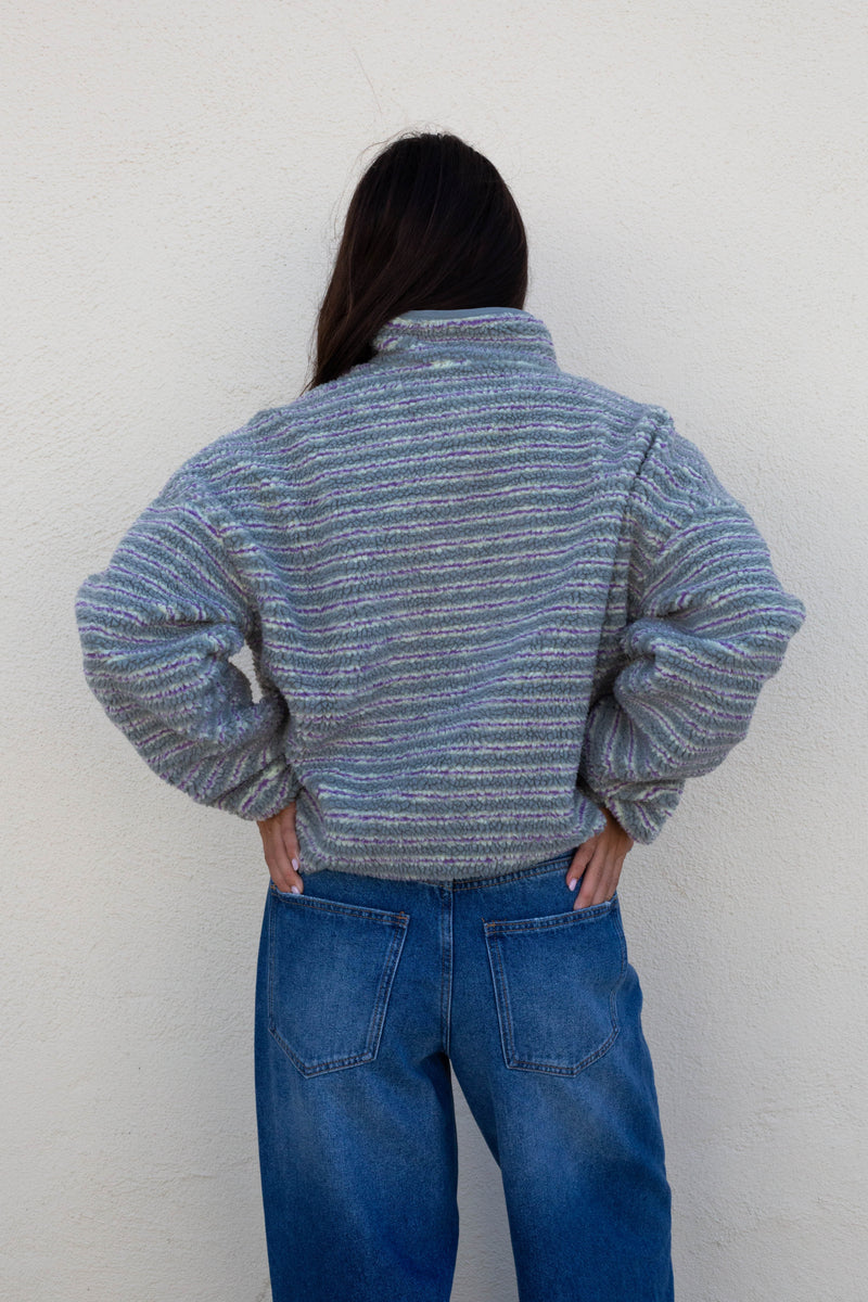 A person shown from behind wearing a gray and blue striped fuzzy cropped sweater and blue jeans, standing against a plain off-white wall.