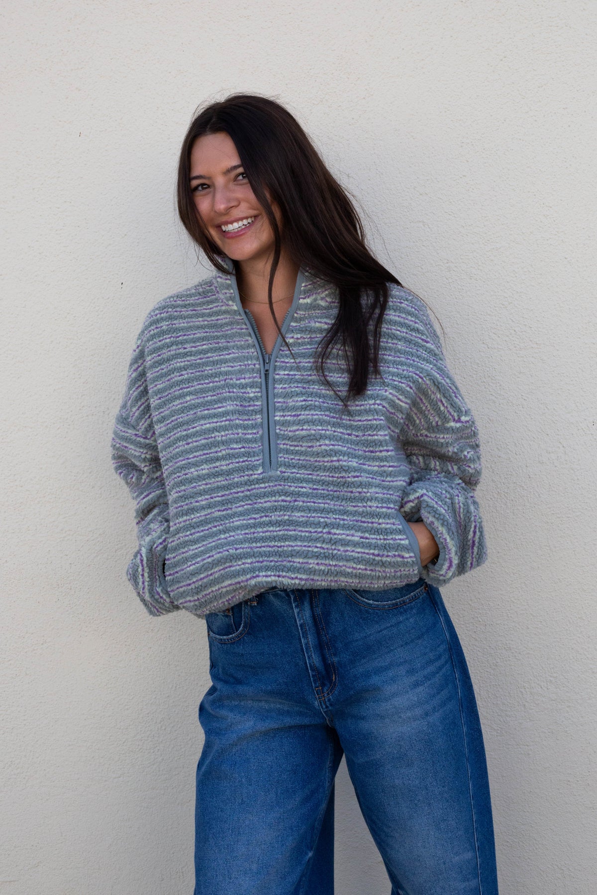 A woman with long dark hair smiling while wearing a gray and blue striped quarter-zip pullover sweater and blue jeans, standing against a plain off-white wall.