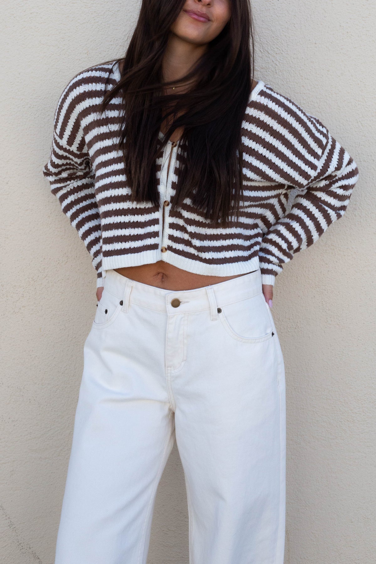 A woman wearing a brown and white striped cropped knit cardigan with a front zipper and drawstring, paired with white wide-leg jeans, standing against a textured beige wall.