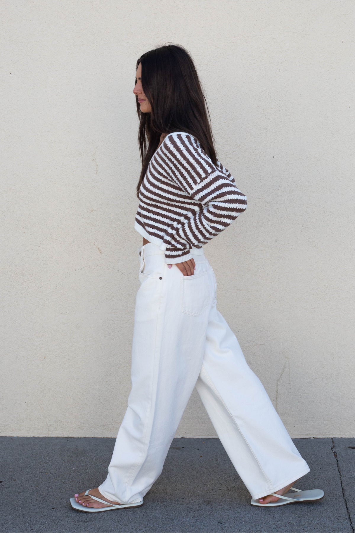A woman in a brown and white striped long-sleeve knit top and white wide-leg jeans walking in profile against a plain beige wall.