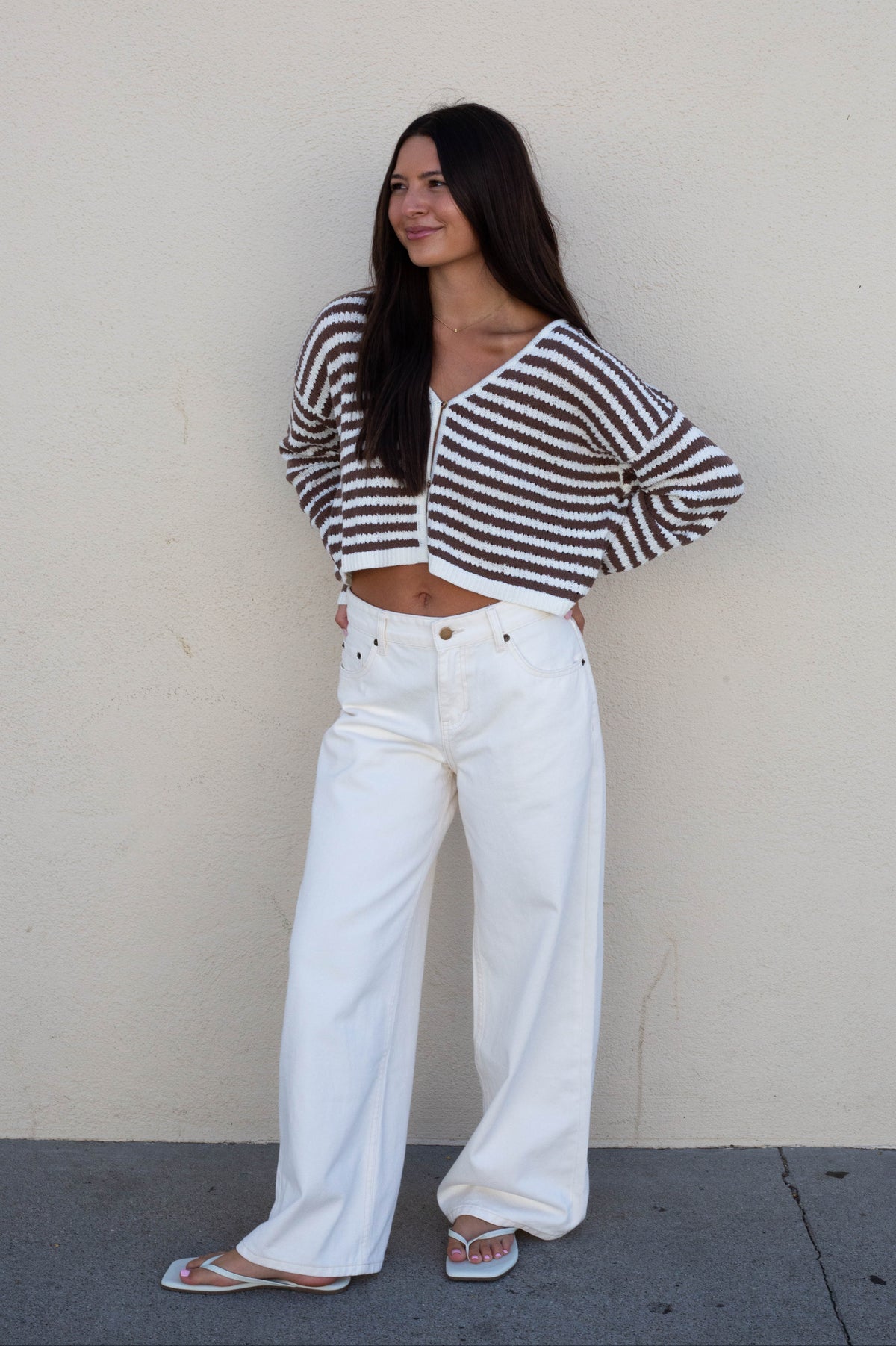 A woman stands against a plain beige wall wearing a brown and white striped cropped knit top, white wide-leg jeans, and white sandals.