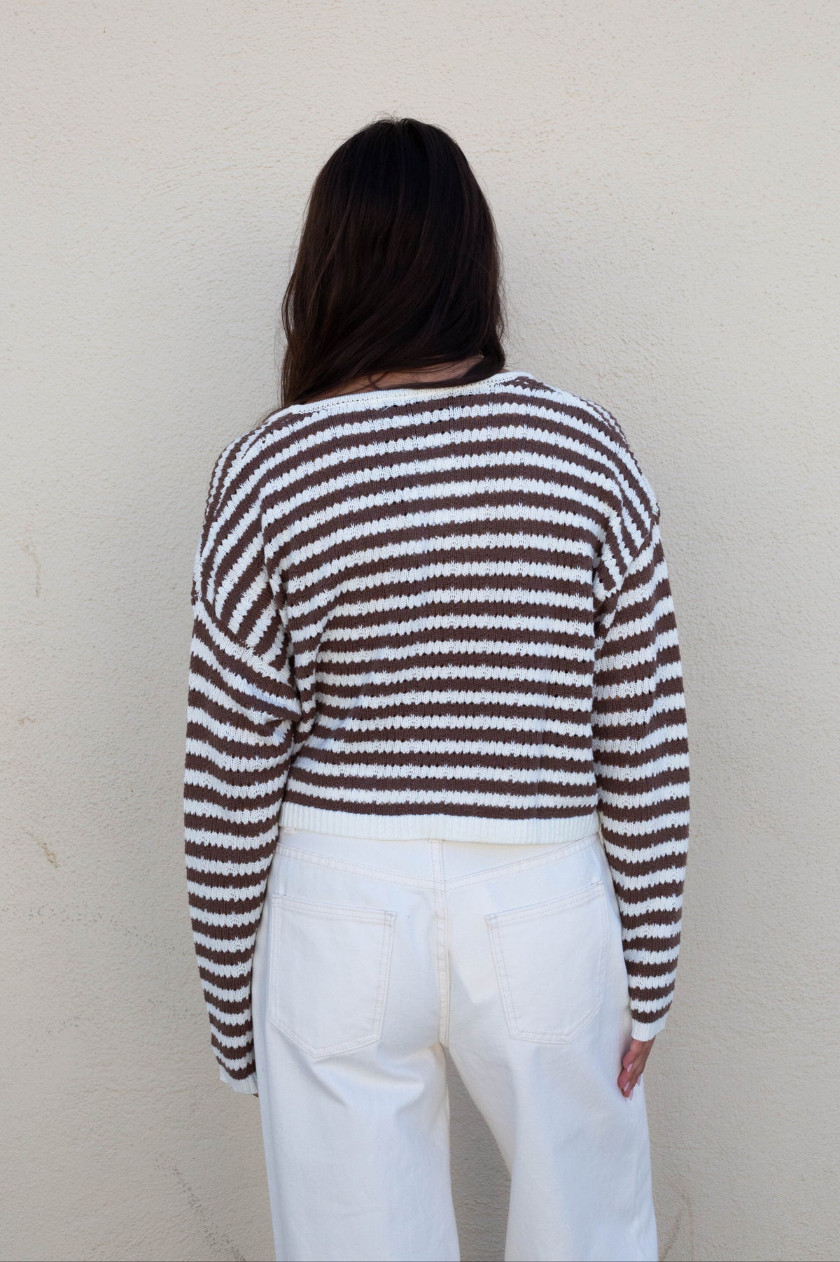 A person viewed from behind wearing a brown and white horizontally striped knit sweater and white pants, standing against a plain off-white wall.