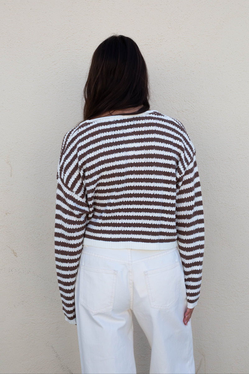 A person viewed from behind wearing a brown and white horizontally striped knit sweater and white pants, standing against a plain off-white wall.