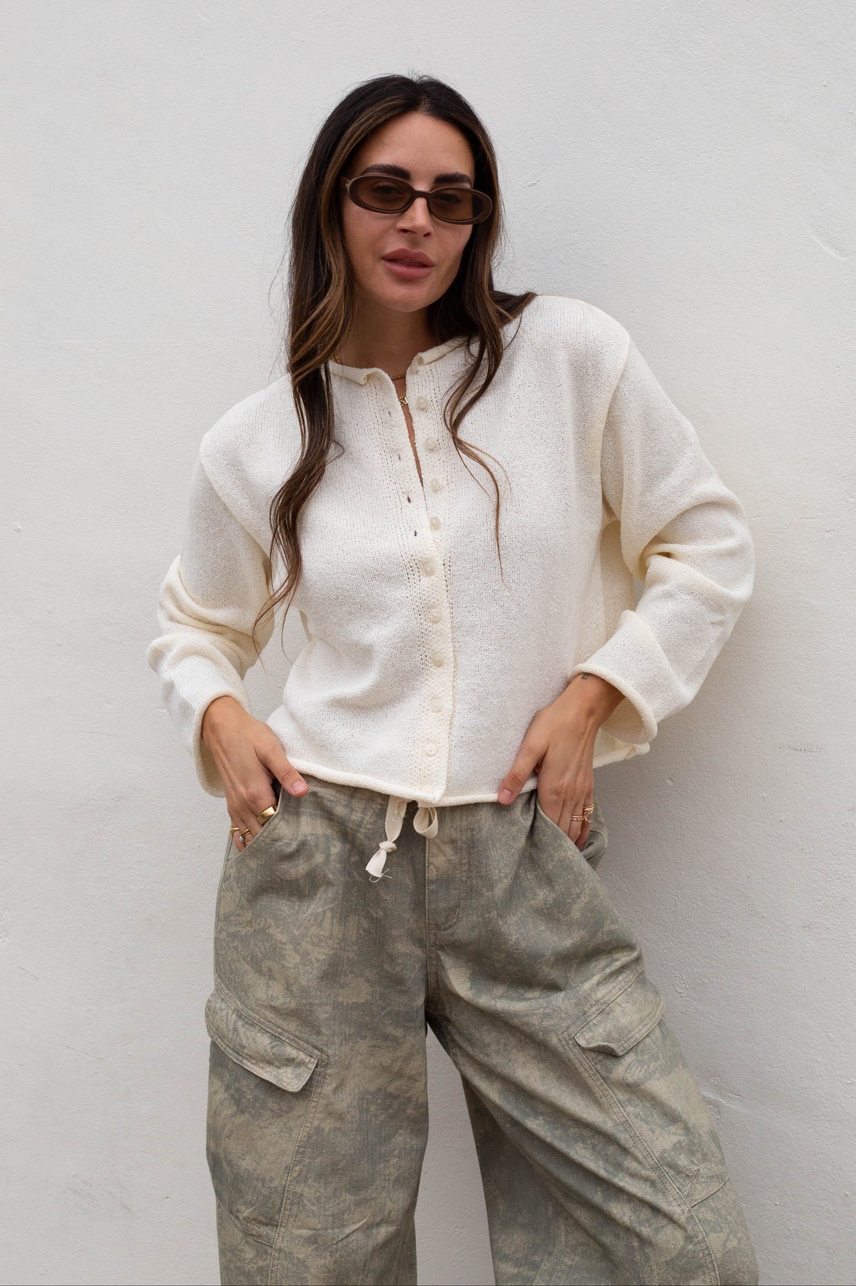 A woman wearing a cream-colored, textured long-sleeve button-up shirt and camouflage cargo pants, standing against a plain white wall.