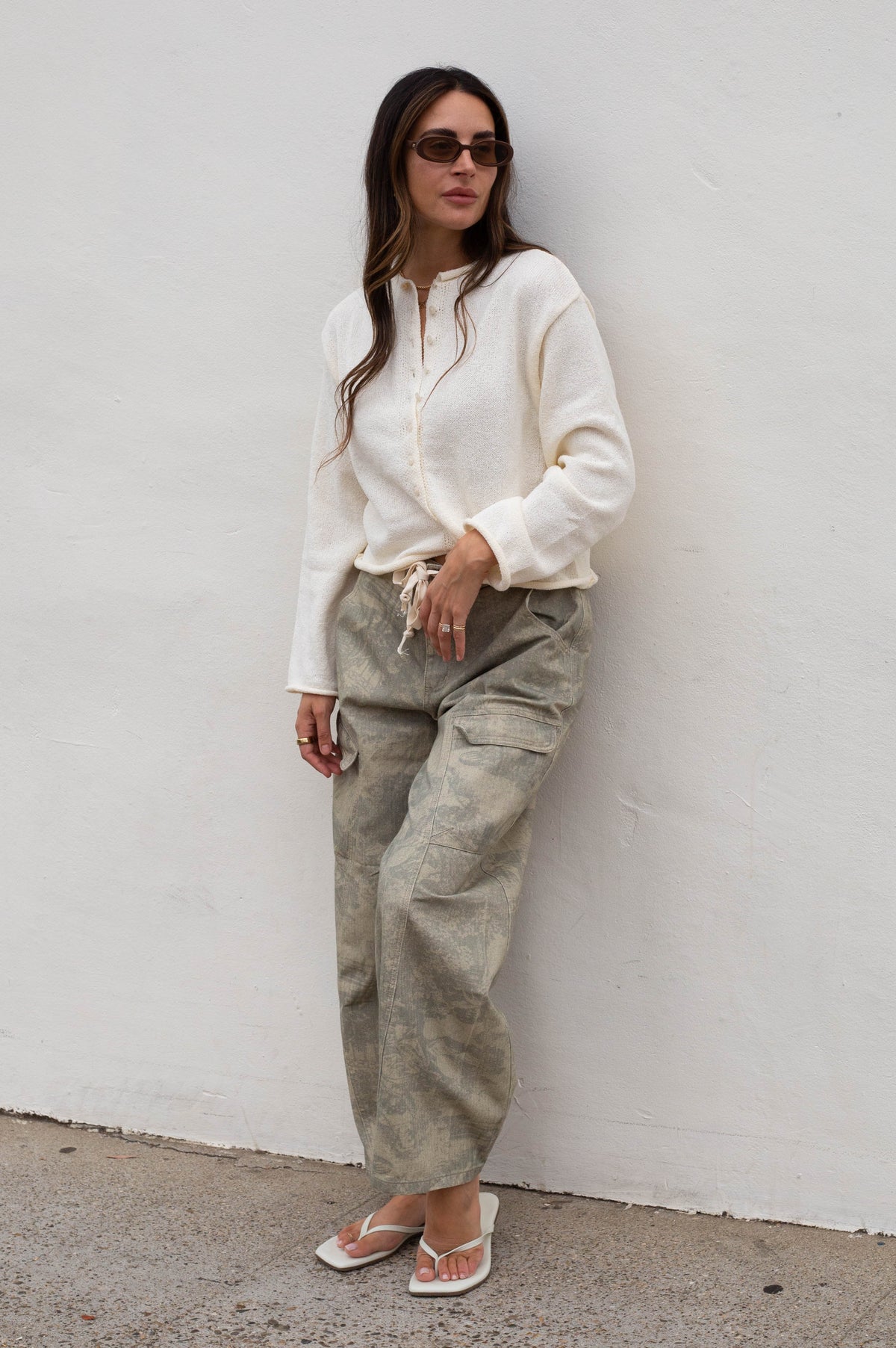 A woman wearing a cream long-sleeve knit top, olive green wide-leg cargo pants, and white sandals stands against a white wall.