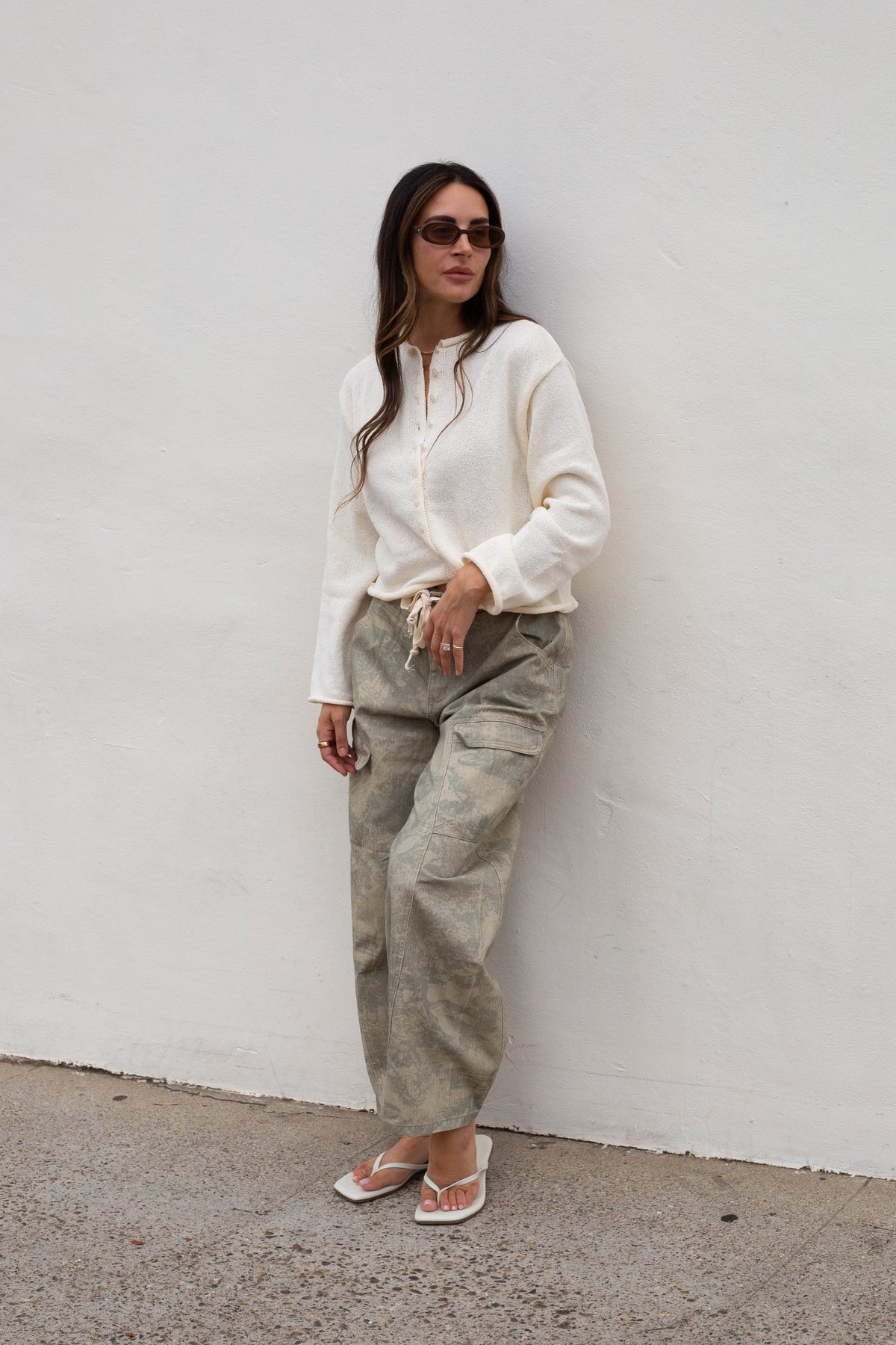 A woman wearing a cream long-sleeve knit top, olive green wide-leg cargo pants, and white sandals stands against a white wall.