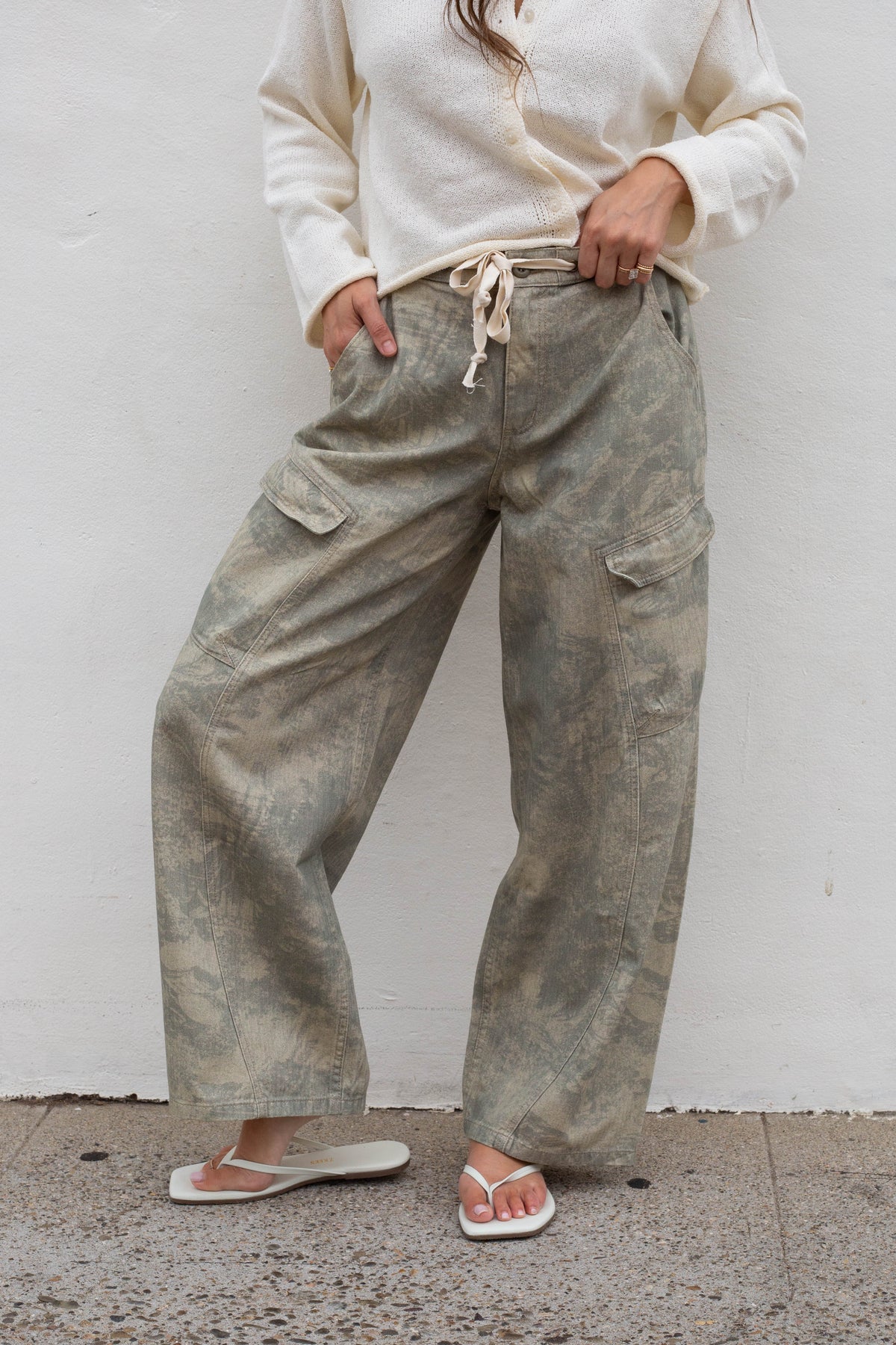 A person wearing loose, wide-leg cargo pants in a muted sage green and gray camouflage pattern, paired with a cream cardigan and white flip-flops, standing against a white wall.