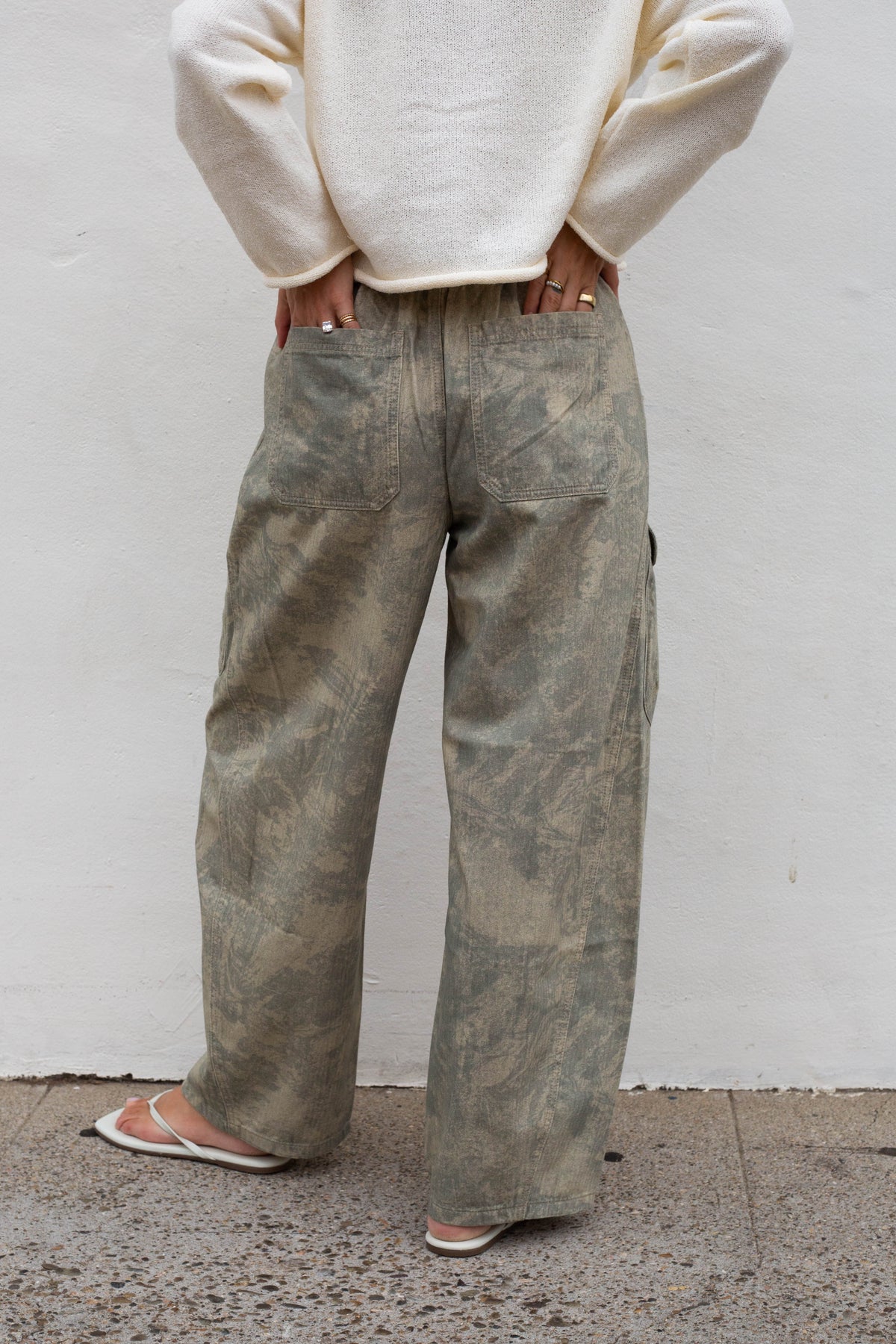 A person shown from the back wearing wide-leg cargo pants with a distressed, faded olive-brown pattern, paired with a cream-colored sweater and white sandals, standing against a white wall.