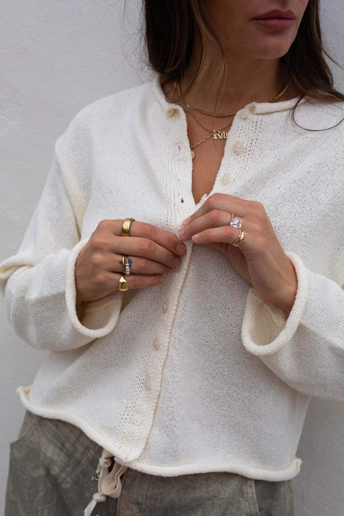A close-up of a woman wearing a cream-colored, textured knit cardigan with buttons, paired with gray pants and gold rings, against a neutral white wall.