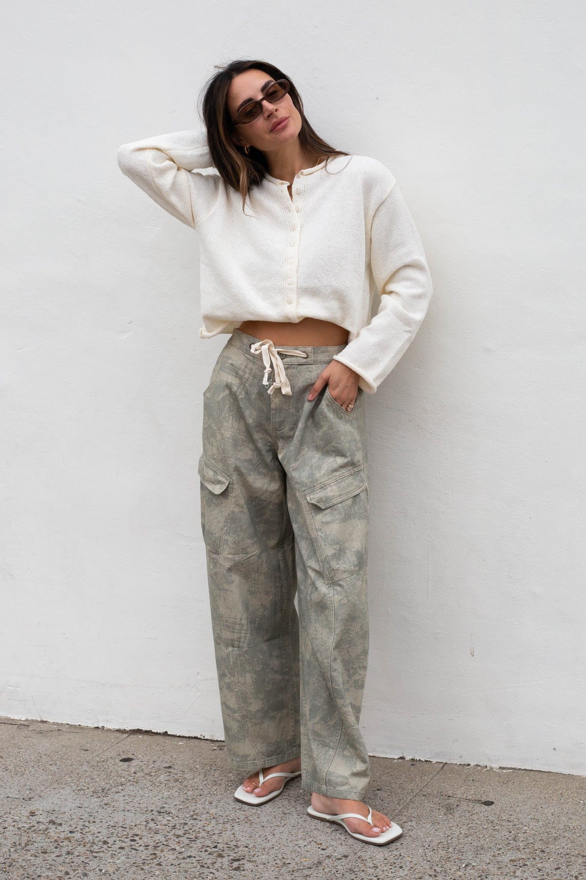 A woman stands against a white wall wearing a cream cropped knit top, loose faded gray-green cargo pants, and white sandals.