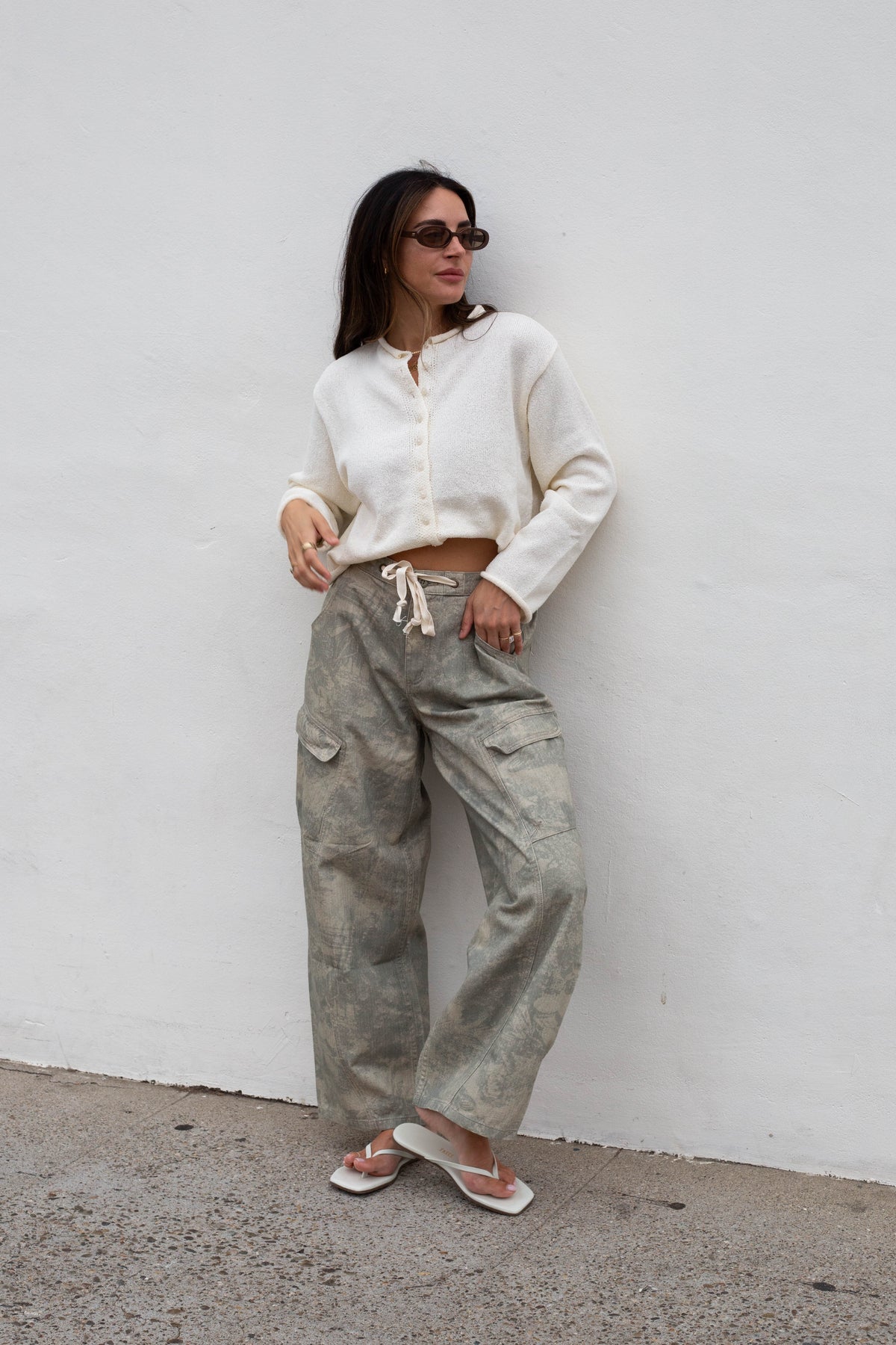 A woman wearing a cream long-sleeve cropped button-up shirt and wide-leg gray cargo pants with a white drawstring, standing against a white wall.