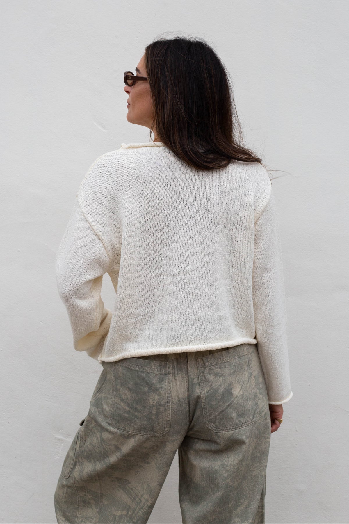 A woman shown from behind wearing a cream-colored, textured knit sweater and gray patterned trousers against a plain white wall.