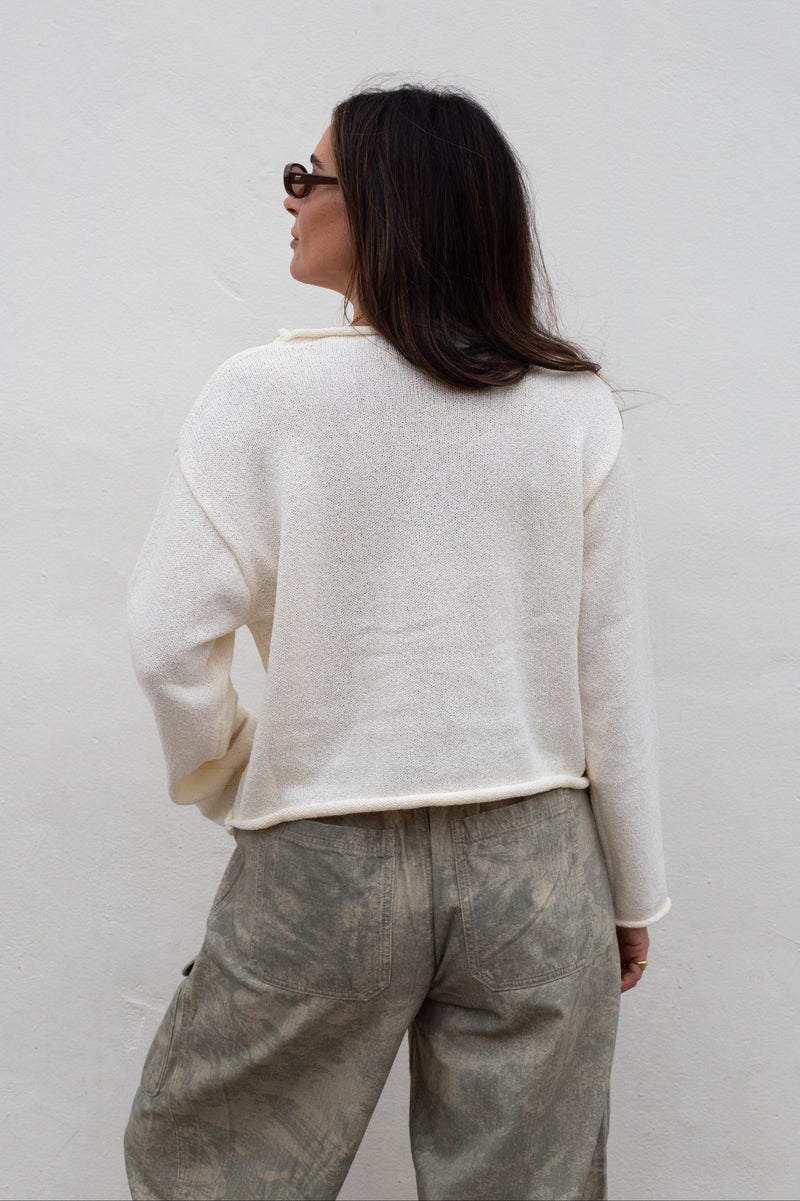 A woman shown from behind wearing a cream-colored, textured knit sweater and gray patterned trousers against a plain white wall.