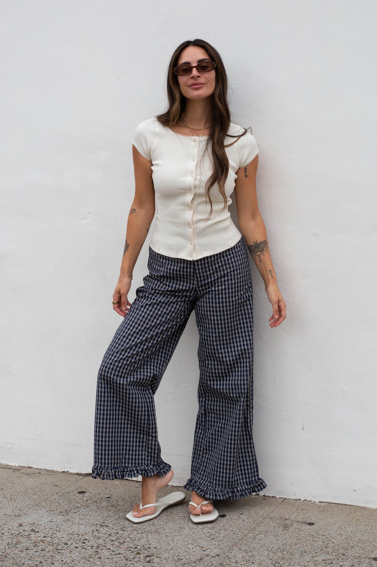 A woman stands against a white wall wearing a cream short-sleeve button-front top and dark navy-and-white gingham wide-leg pants with ruffled hems, paired with white sandals.