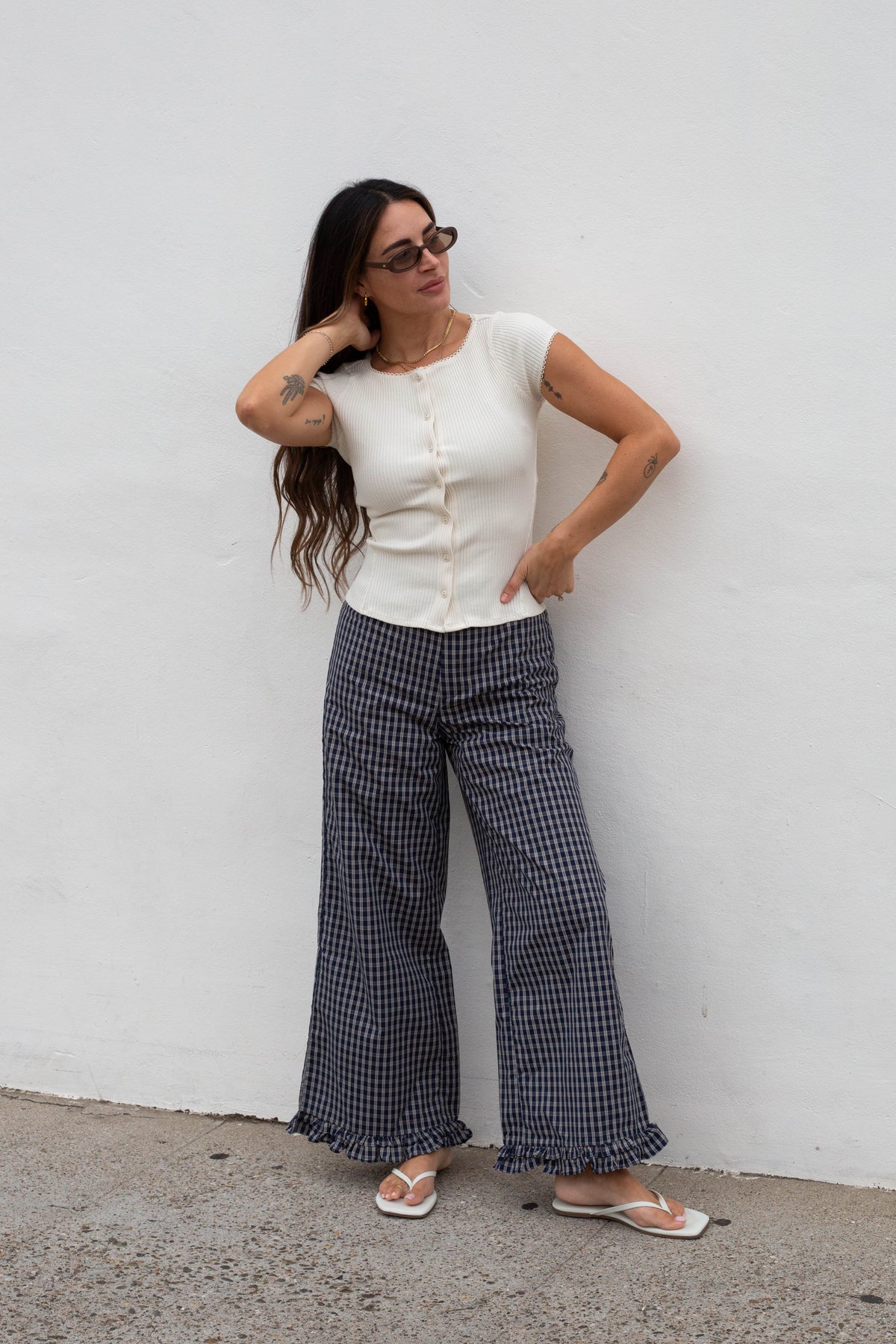 A woman wearing a white short-sleeve button-up top and navy and white gingham wide-leg pants, standing against a white wall.