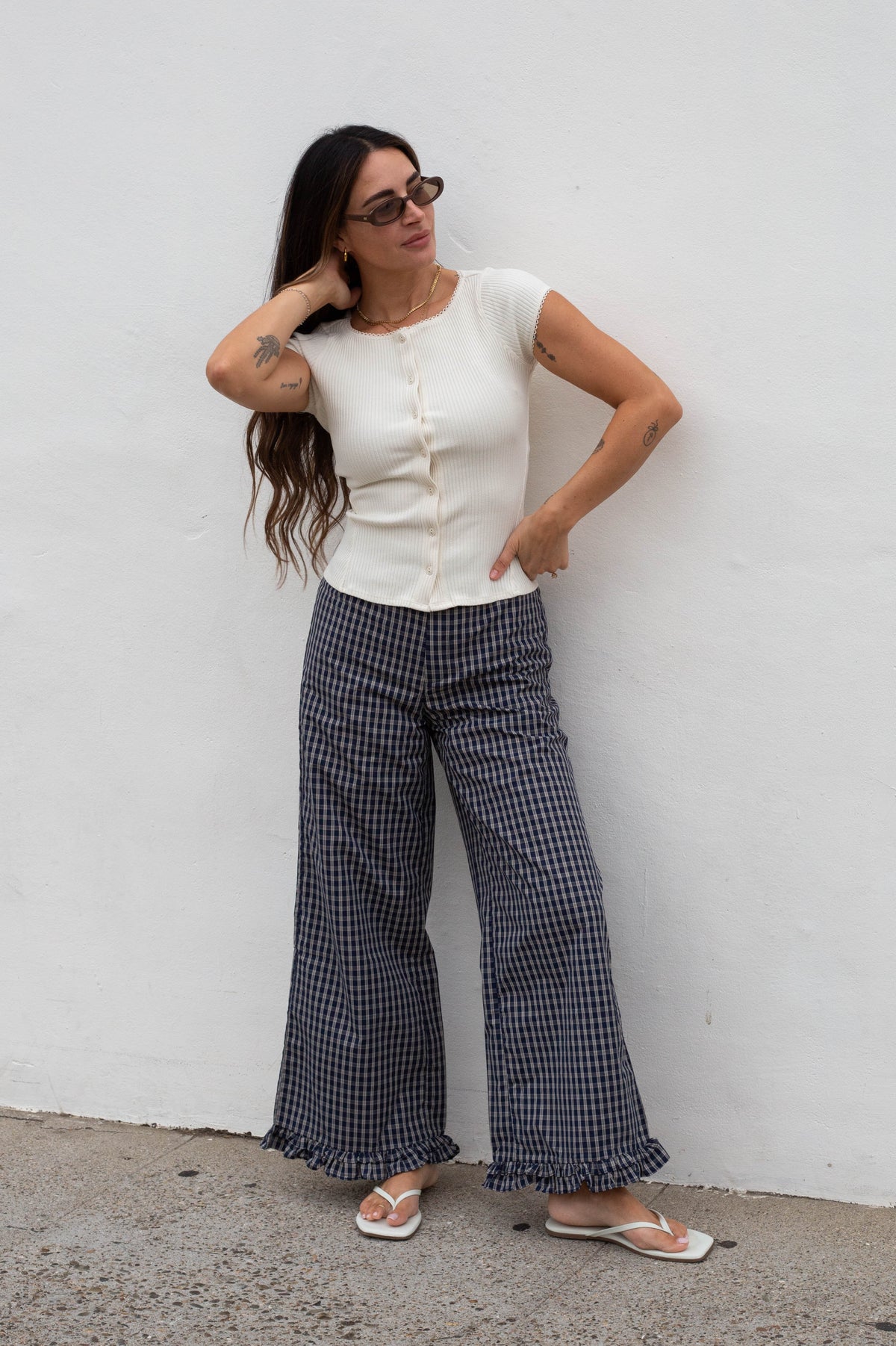 A woman wearing a white short-sleeve button-up top and navy and white gingham wide-leg pants, standing against a white wall.