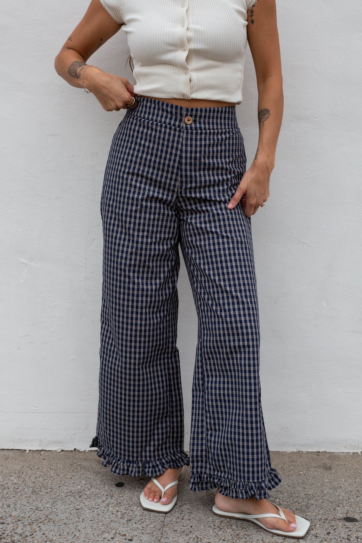 A person wearing navy and white gingham wide-leg cropped pants with a ruffled hem, paired with a cream ribbed crop top and white flip-flops, standing against a white wall.