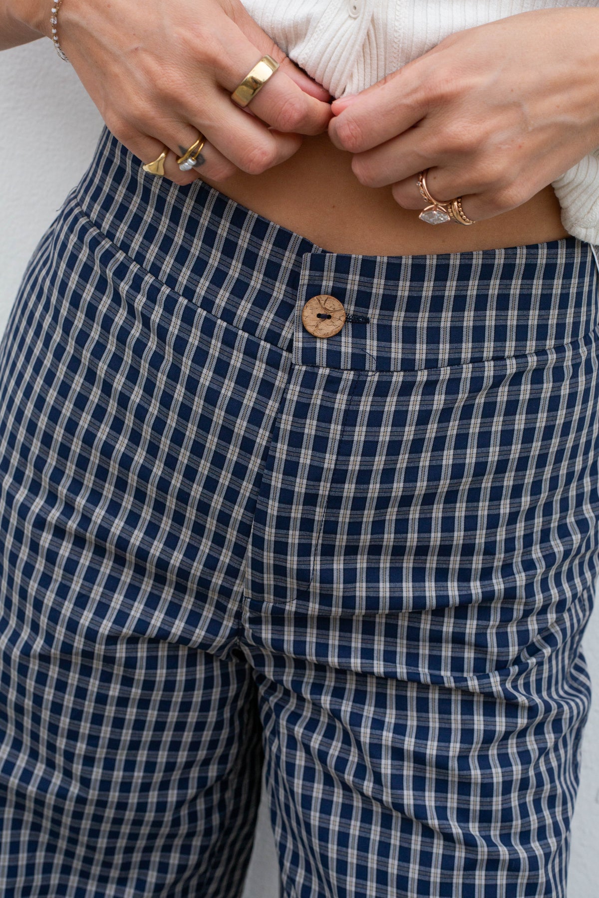 Close-up of a person wearing navy and white gingham check pants, pulling the hem up with both hands. The pants feature a single tan button and a blue-and-white checkered pattern.