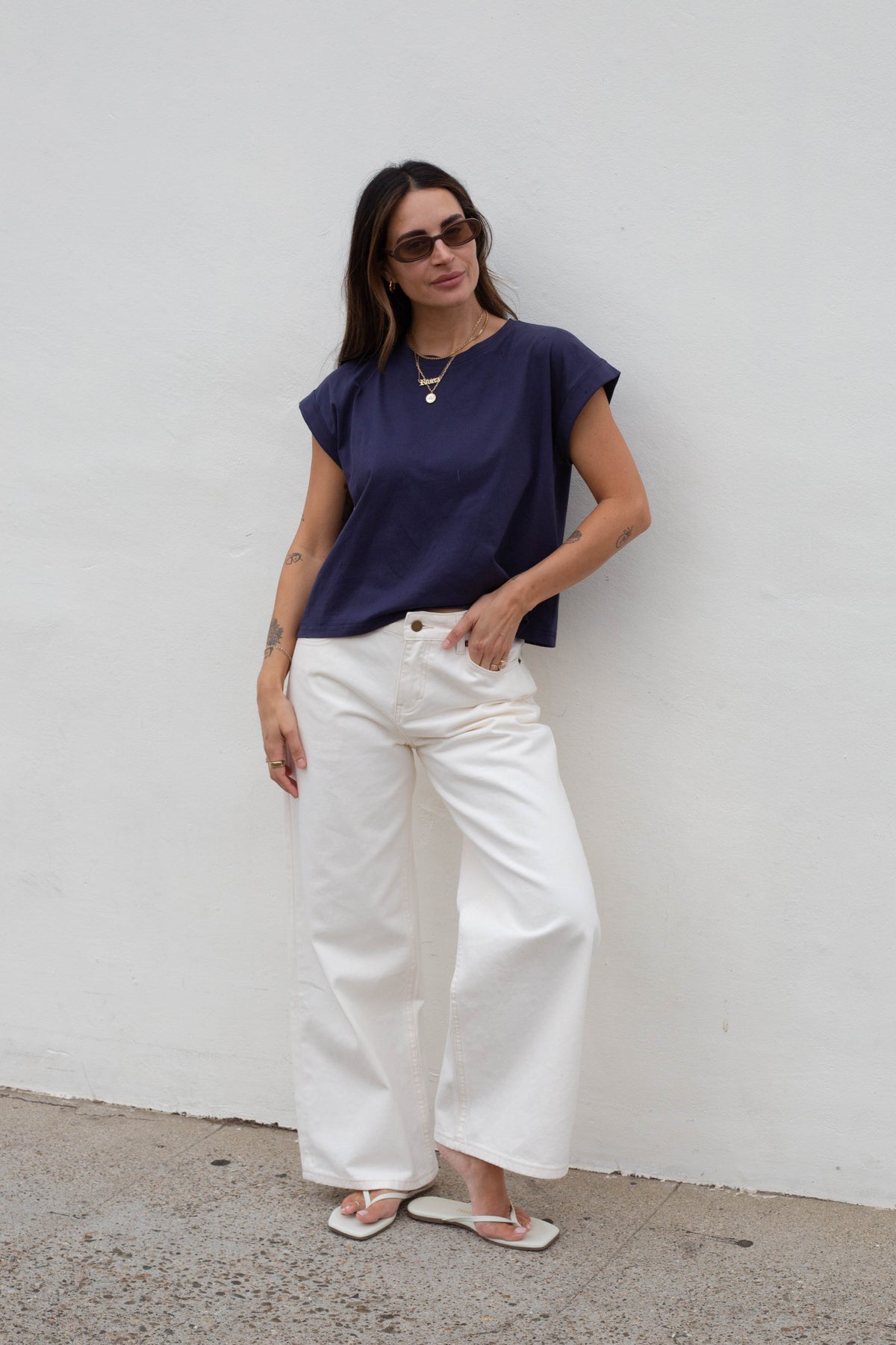 A woman stands against a white wall wearing a navy blue short-sleeve top, white wide-leg pants, and white sandals.