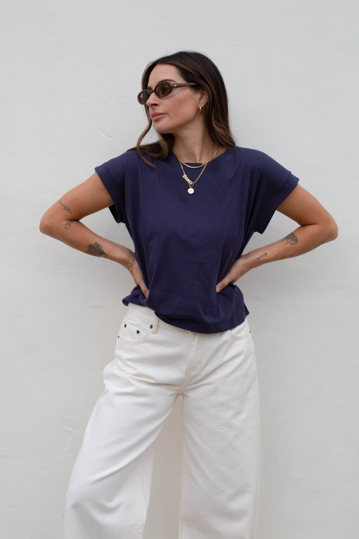 A woman wearing a navy blue short-sleeve t-shirt and white wide-leg pants, posing against a plain white wall.