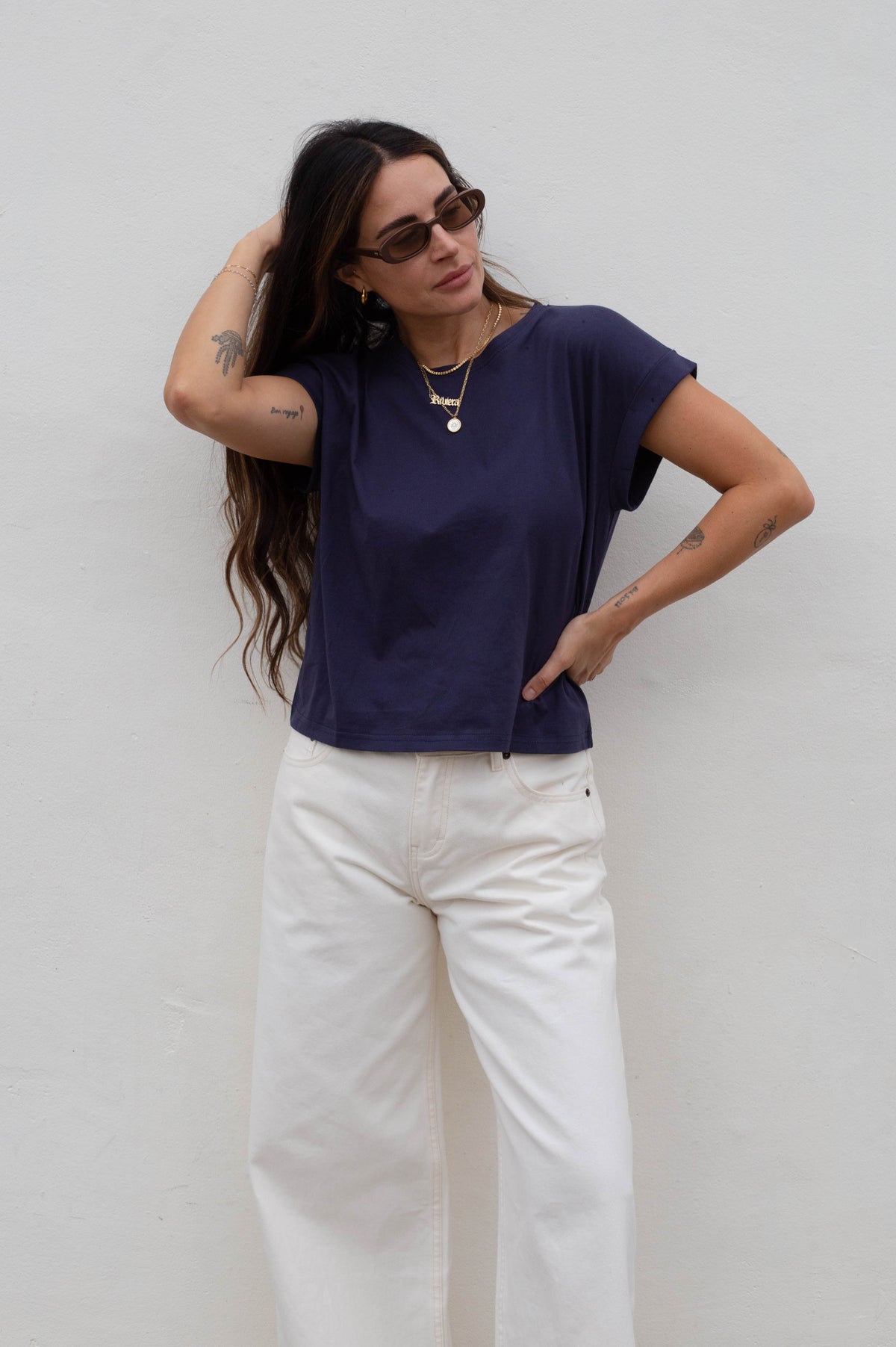 A woman wearing a navy blue short-sleeve t-shirt and white wide-leg jeans, posing against a plain white wall.