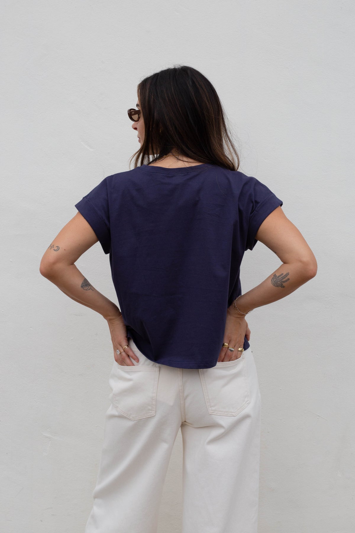 A woman viewed from behind wearing a dark navy blue short-sleeve t-shirt and off-white wide-leg trousers, standing against a plain light gray wall.