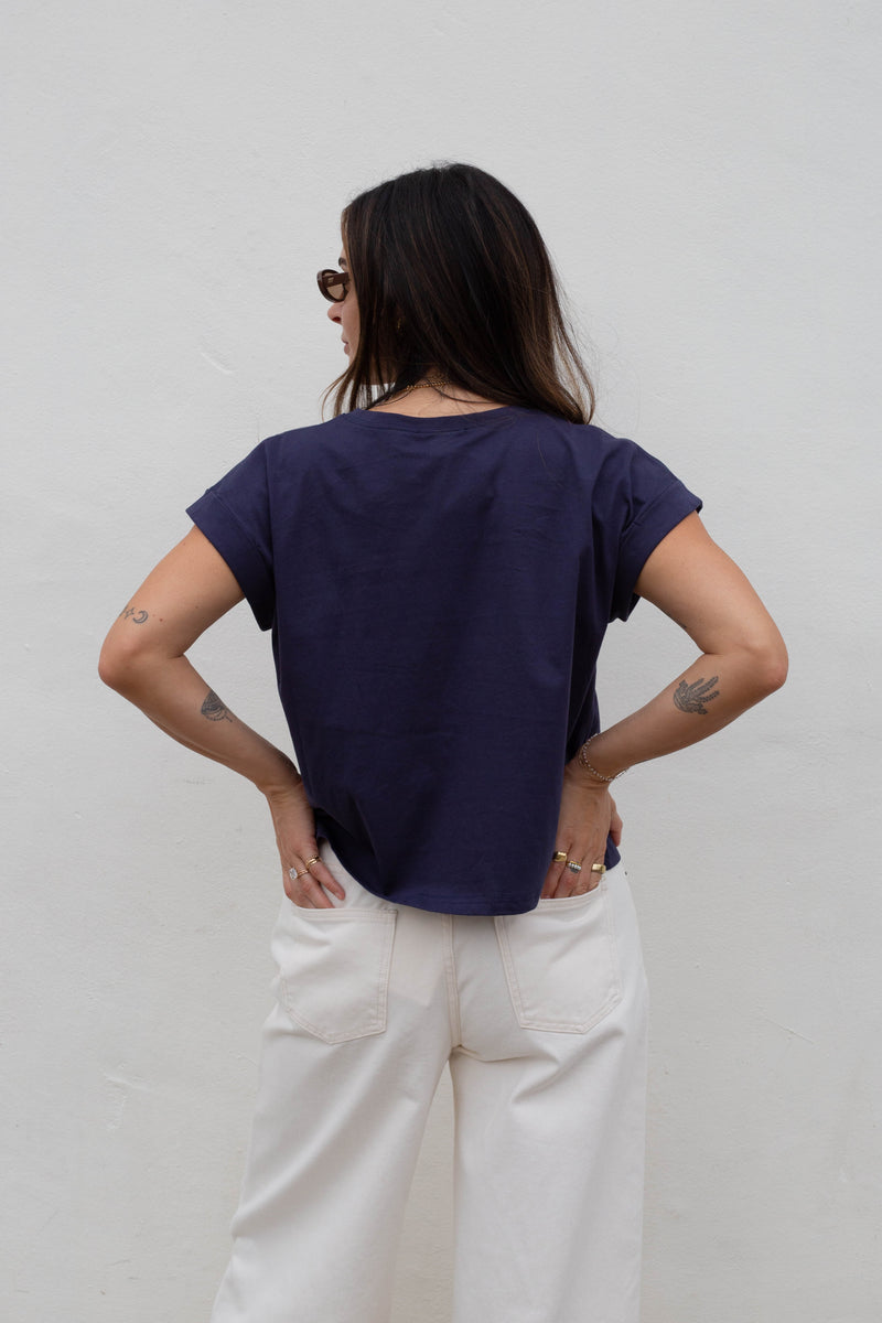 A woman viewed from behind wearing a dark navy blue short-sleeve t-shirt and off-white wide-leg trousers, standing against a plain light gray wall.