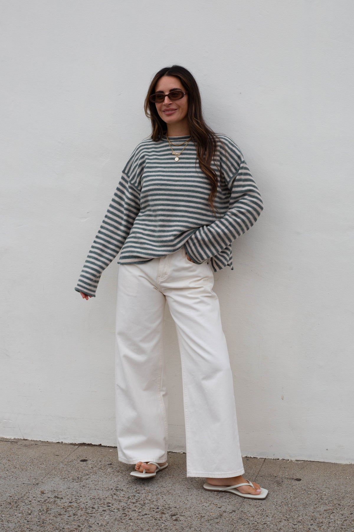 A woman wearing a green and white striped long-sleeve top, white wide-leg pants, and sunglasses, standing against a plain white wall.