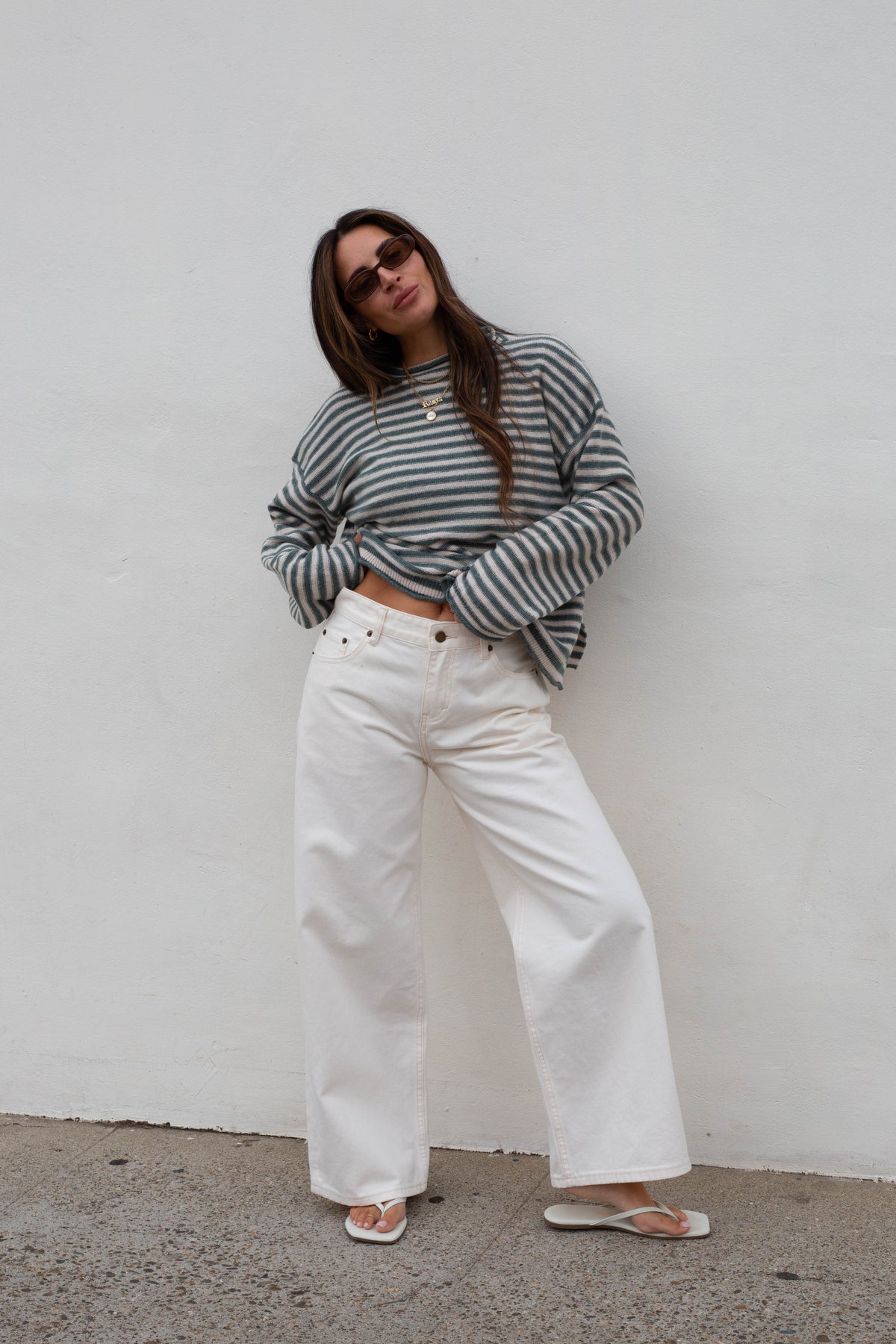 A woman wearing a gray and white striped long-sleeve crop top, white wide-leg jeans, and white flip-flops, standing against a plain white wall.