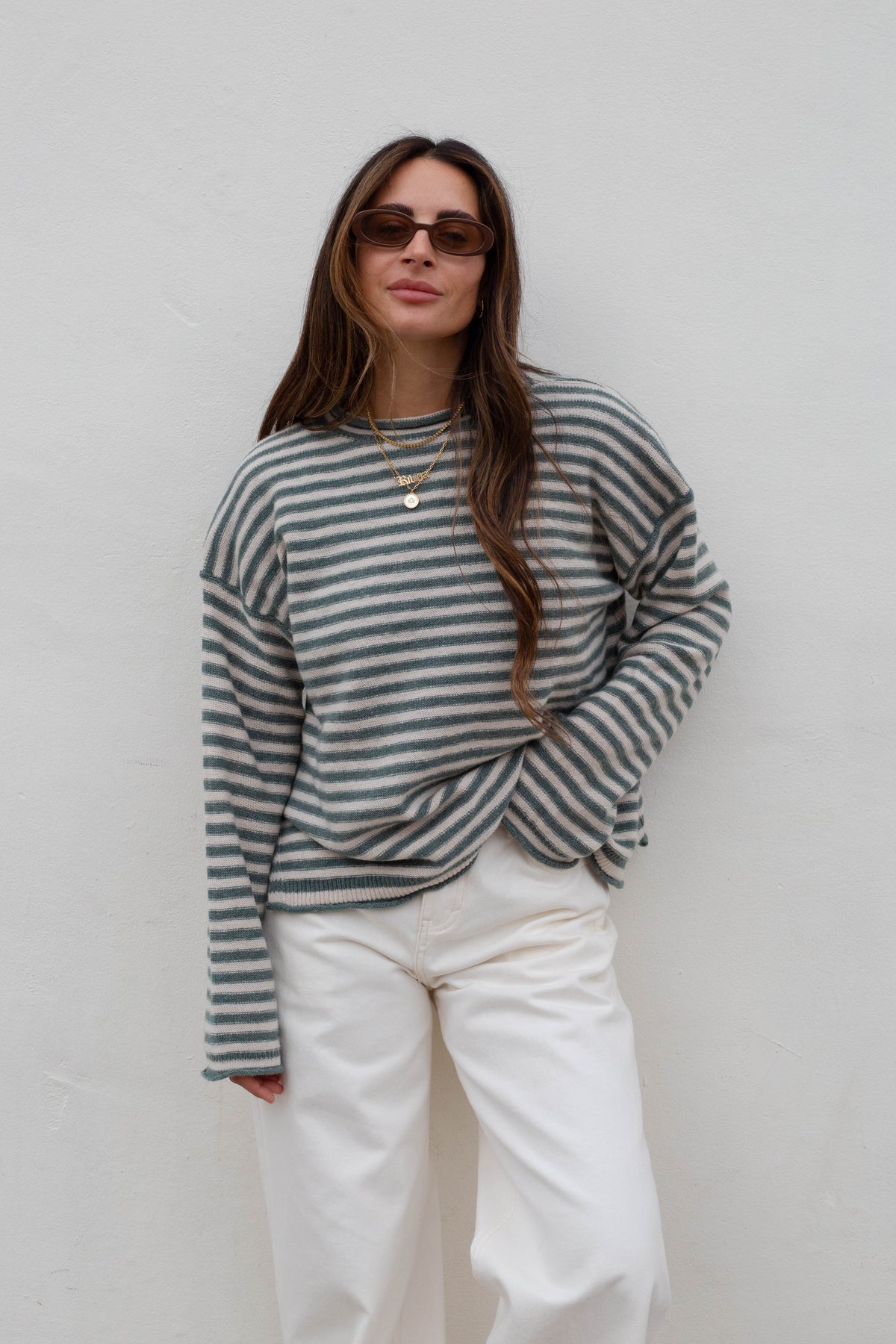A woman wearing a gray and white horizontally striped long-sleeve knit top, white pants, and sunglasses, standing against a plain off-white wall.