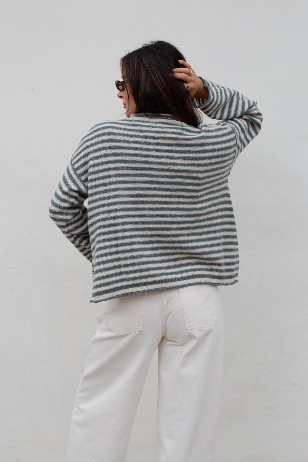 A woman viewed from behind wearing a striped knit sweater with white pants, standing against a plain light gray wall.