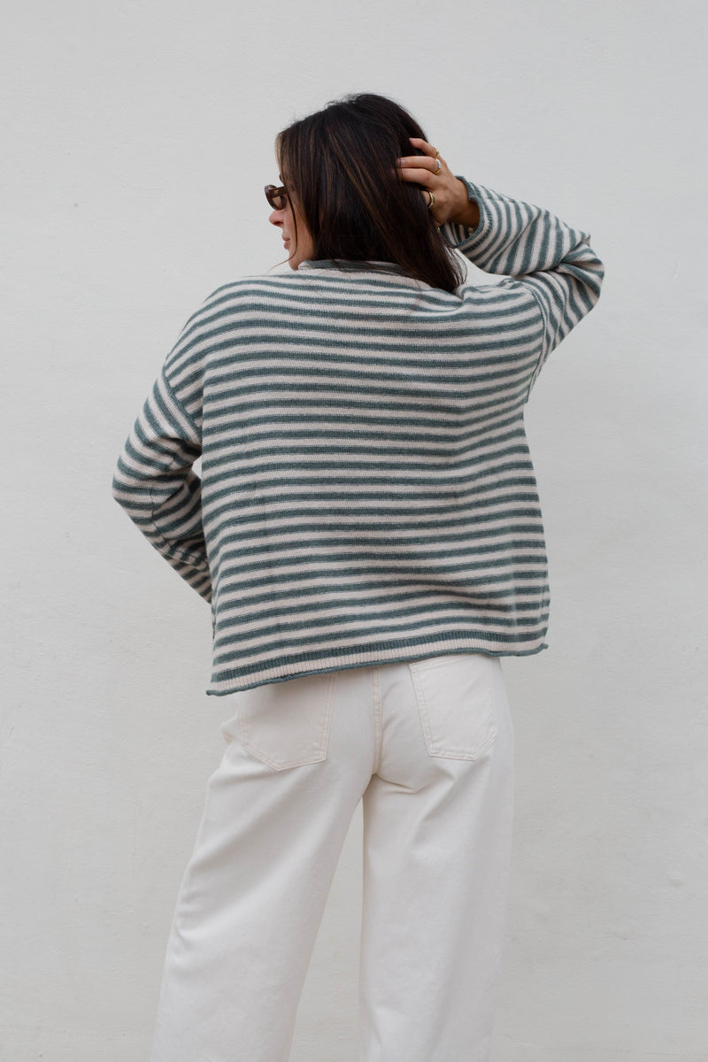 A woman viewed from behind wearing a striped knit sweater with white pants, standing against a plain light gray wall.