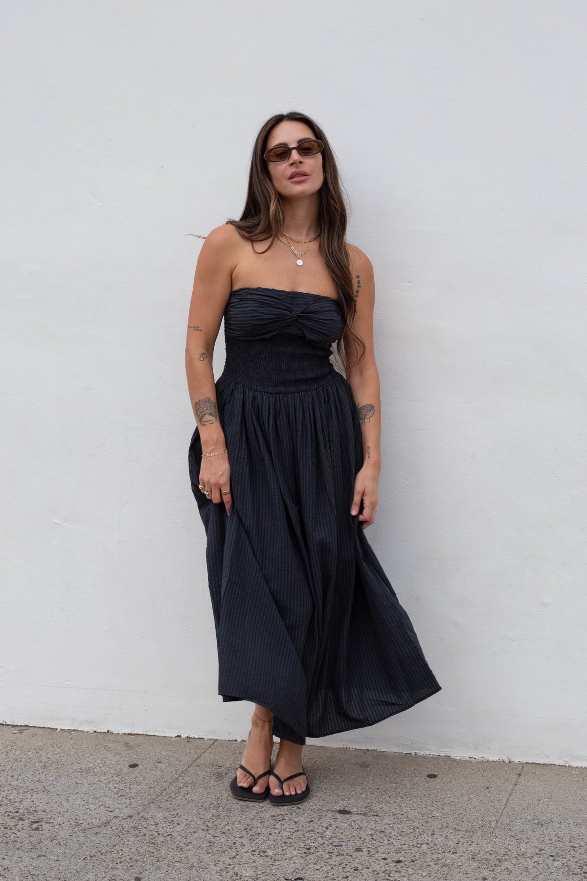 A woman wearing a strapless black midi dress with a gathered waist and flowing skirt, standing against a plain white wall.