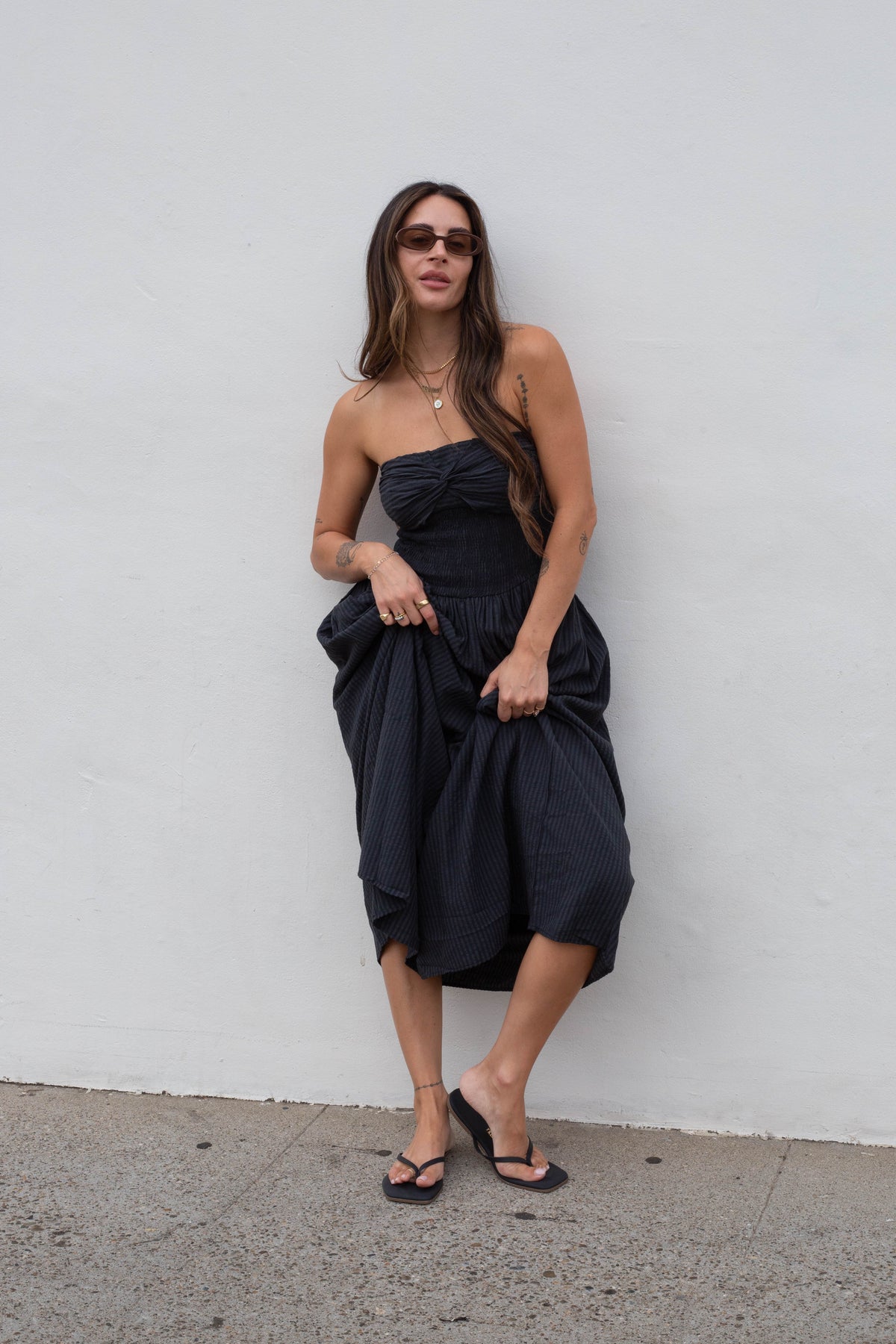 A woman wearing a black strapless midi dress with a gathered waist and voluminous skirt, standing against a plain white wall.