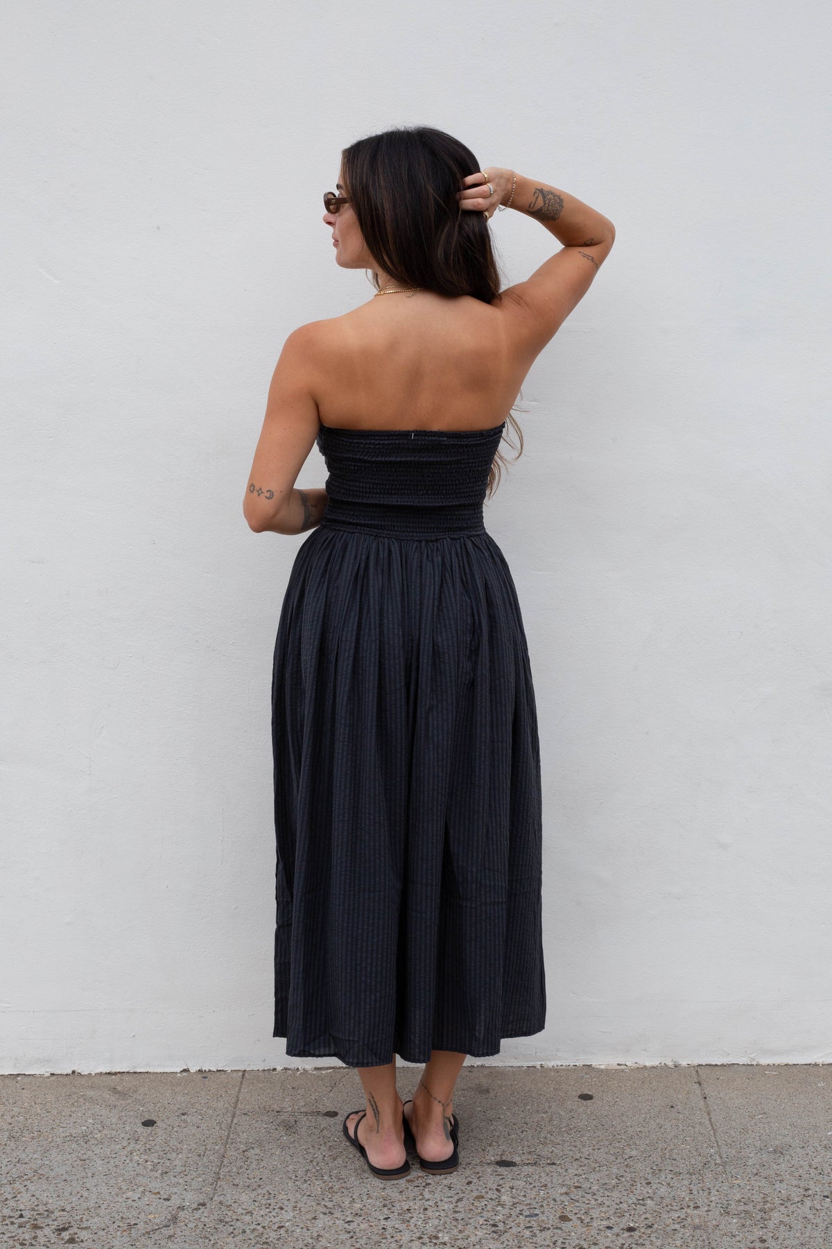A woman stands with her back to the camera wearing a strapless black midi dress with a pleated skirt, set against a plain white wall.