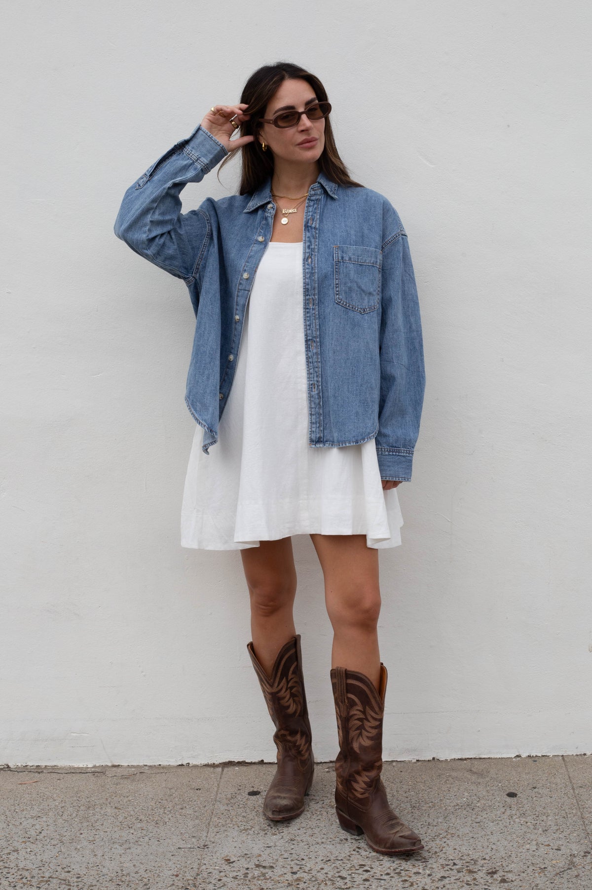 A woman stands against a white wall wearing a light blue denim shirt over a white dress, with brown cowboy boots and dark sunglasses.