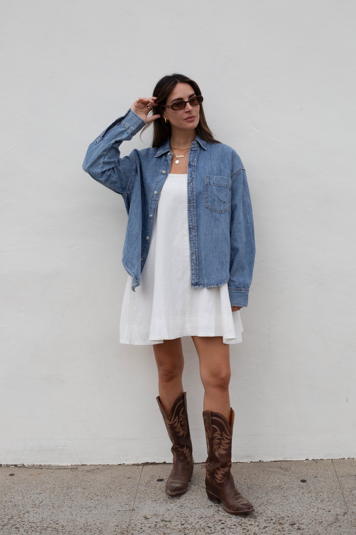 A woman stands against a white wall wearing a light blue denim shirt over a white dress, with brown cowboy boots and dark sunglasses.