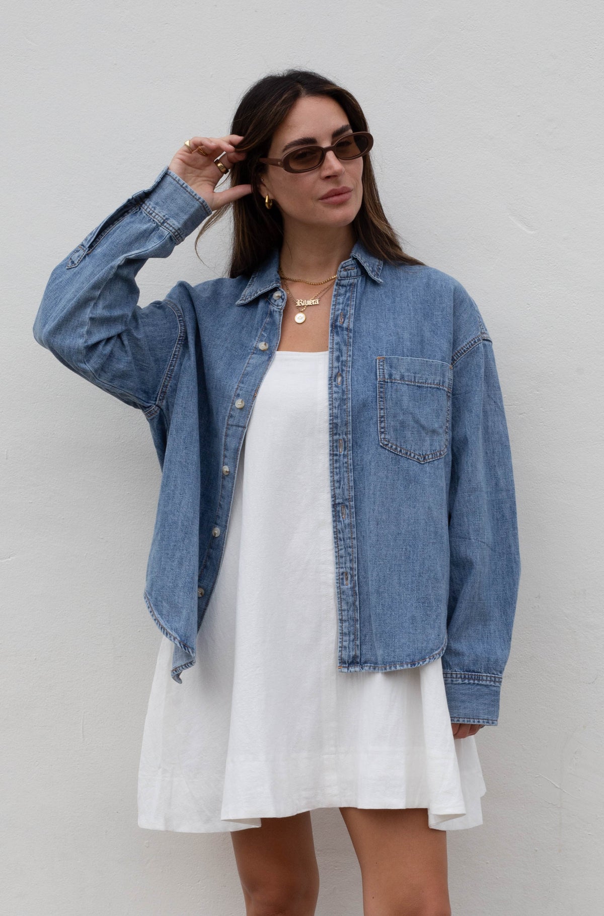A woman stands against a white wall wearing a light blue denim shirt over a white dress, with brown cowboy boots and dark sunglasses.