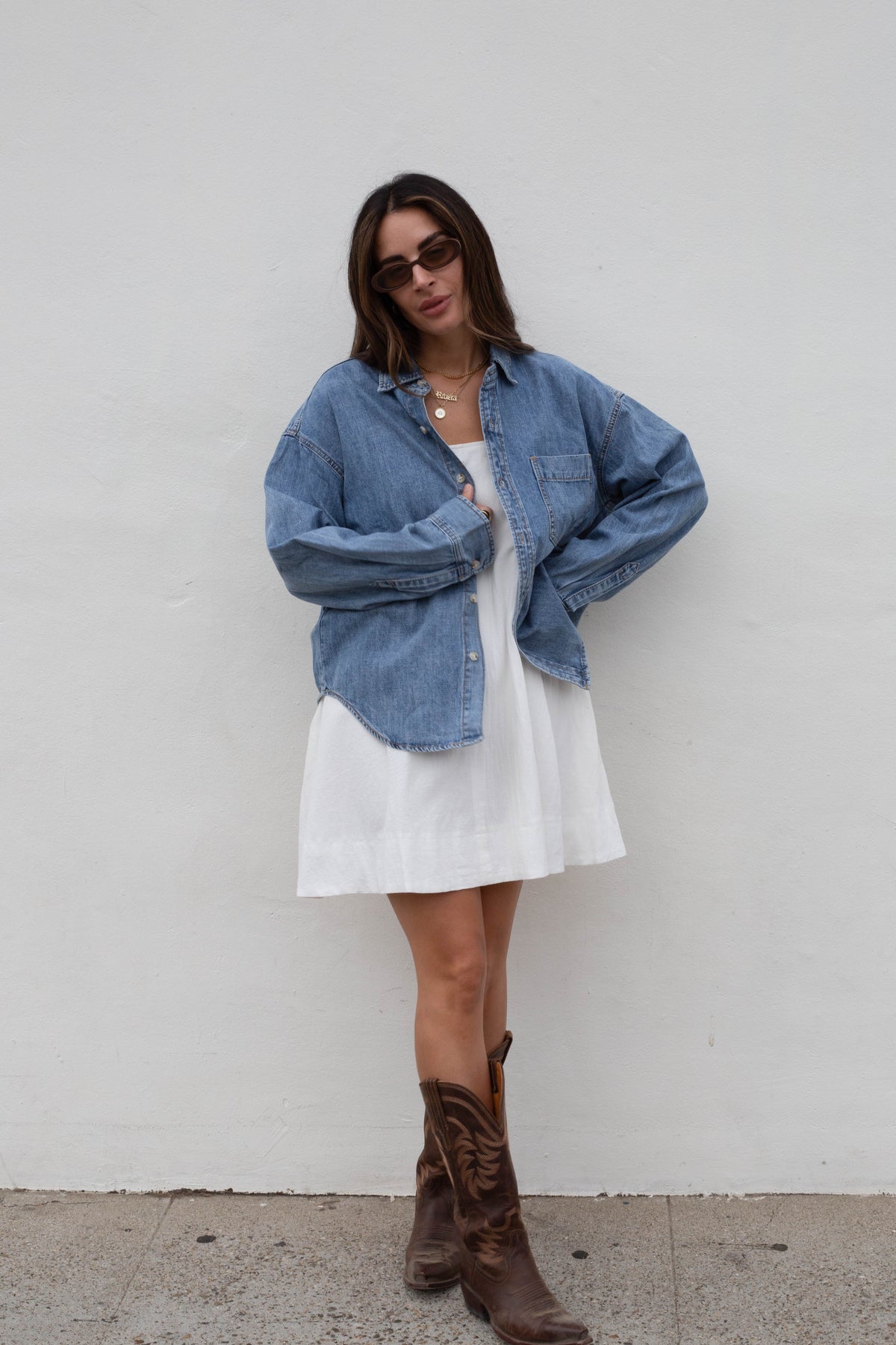 A woman stands against a white wall wearing an oversized light blue denim shirt over a white dress, with brown cowboy boots and dark sunglasses.