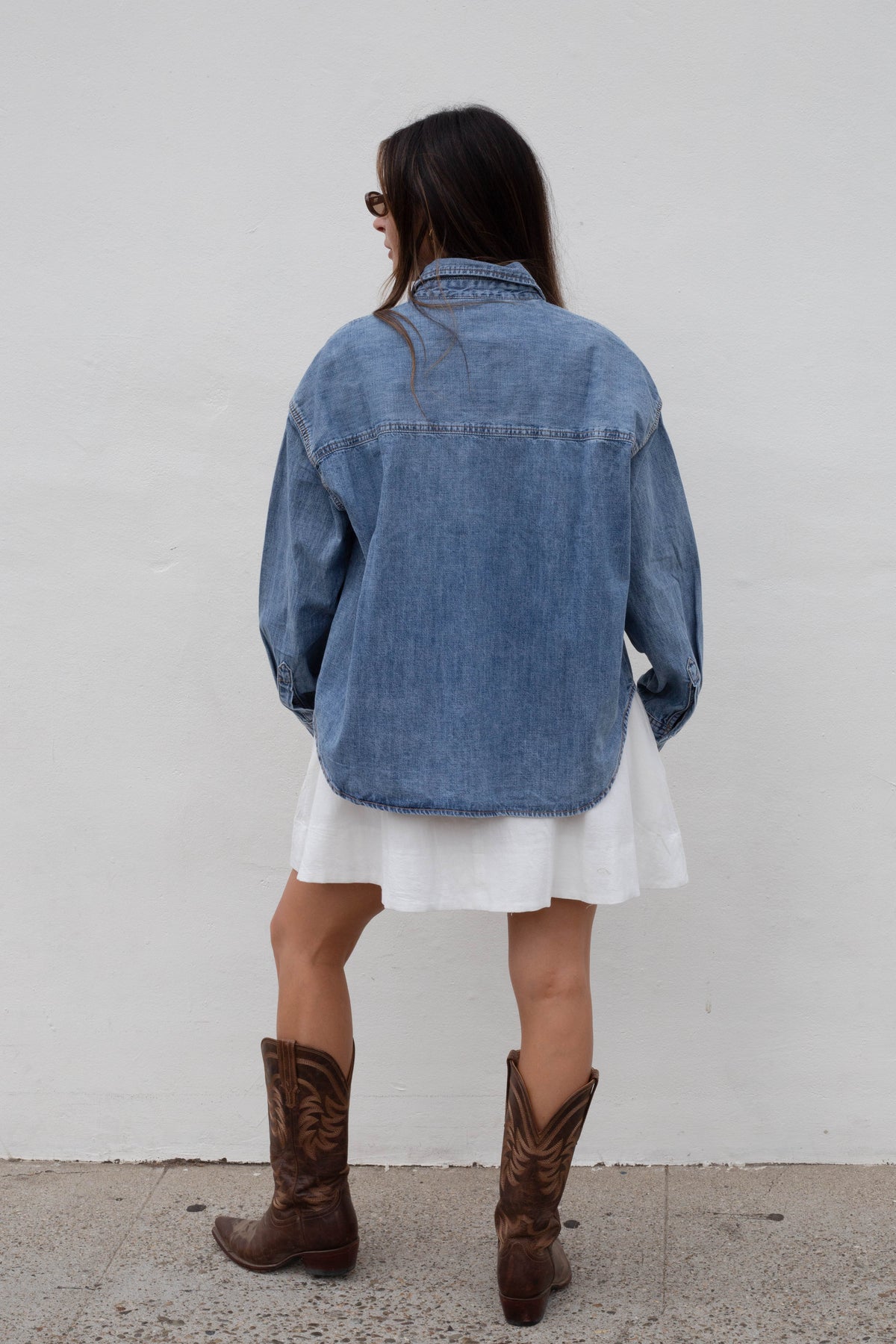 A woman shown from behind wearing an oversized blue denim shirt over a white dress, paired with brown cowboy boots, standing against a plain white wall.